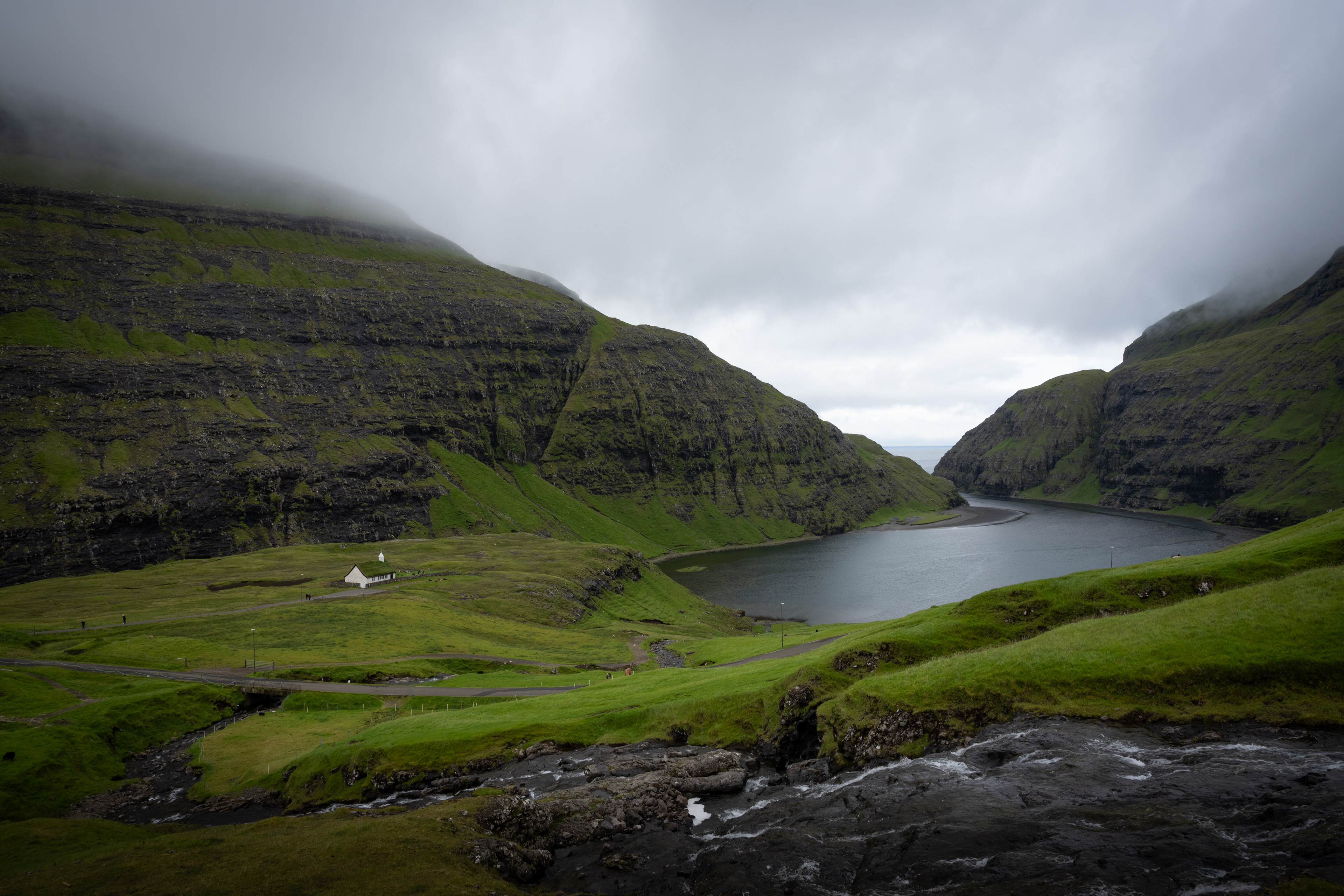 A narrow valley opens toward water under a low ceiling of cloud. The church rests within the land rather than apart from it, shaped by terrain and weather more than intention.