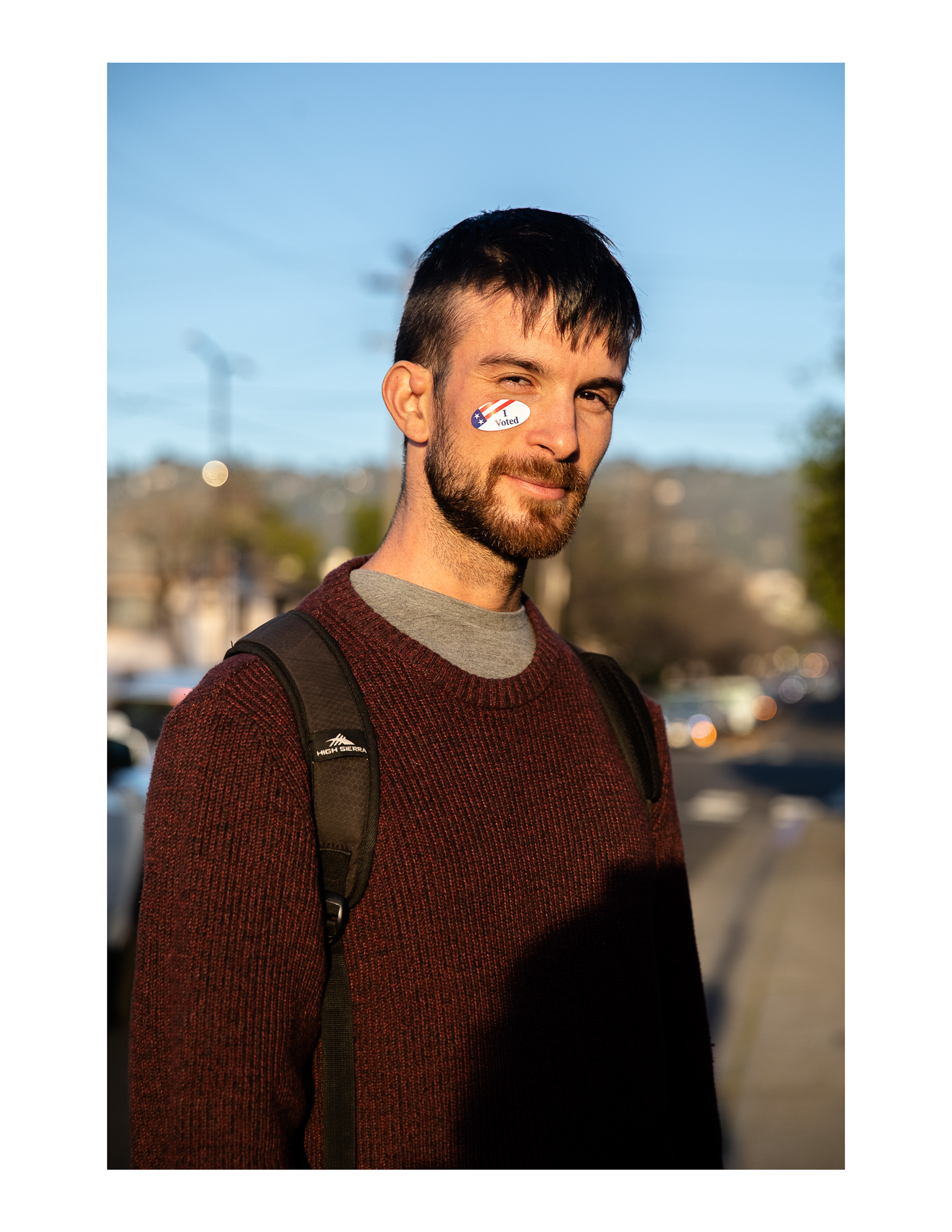 Joseph is wearing a burgundy, tight-cabled sweater over a grey t-shirt, black backpack straps over his shoulders. He has light skin, a brown beard and short dark hair, and is squinting against the late afternoon sunlight into the camera, with his "I Voted" sticker on his right cheek.