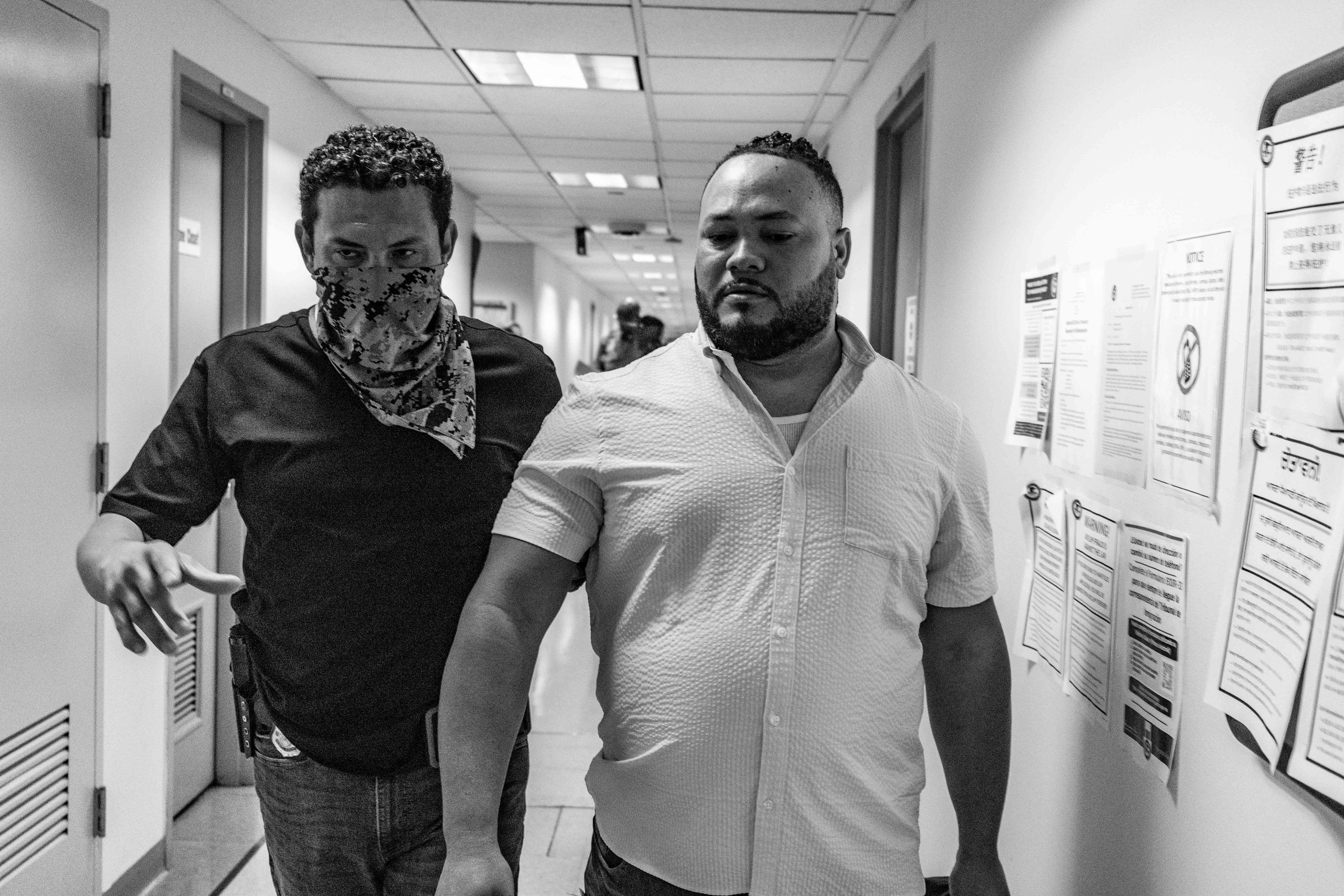 A Customs and Border Patrol officer detains and leads a respondent down the hallway towards the side stairwell.