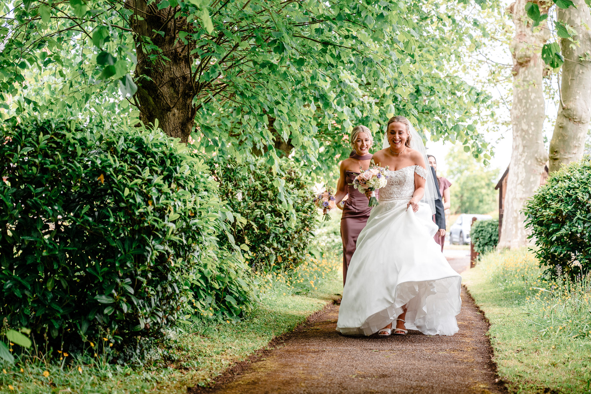 The bride makes her way to the church through a green canopy of tree leaves.