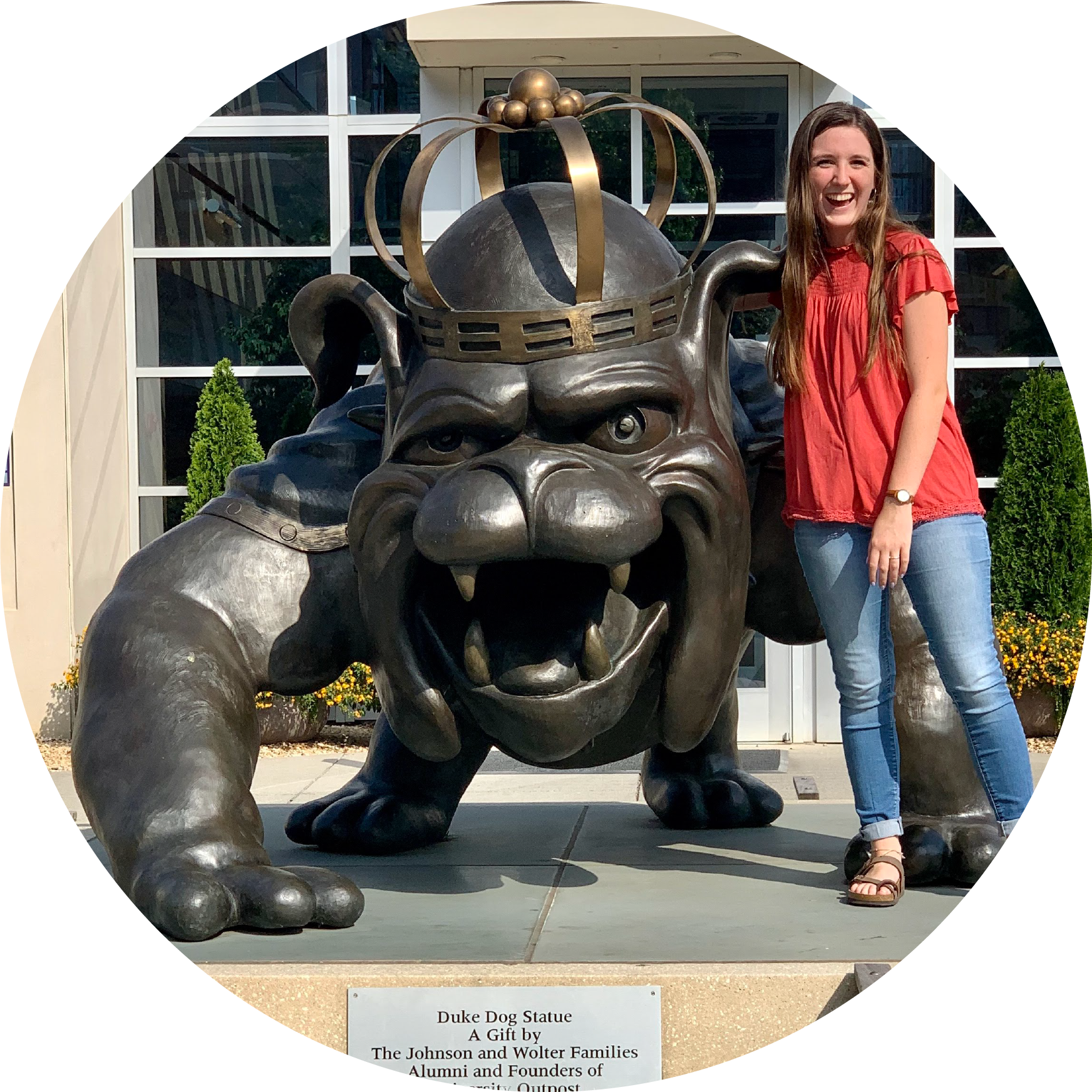 A woman in a red shirt and jeans standing next to a statute of James Madison University's Duke Dog statue.
