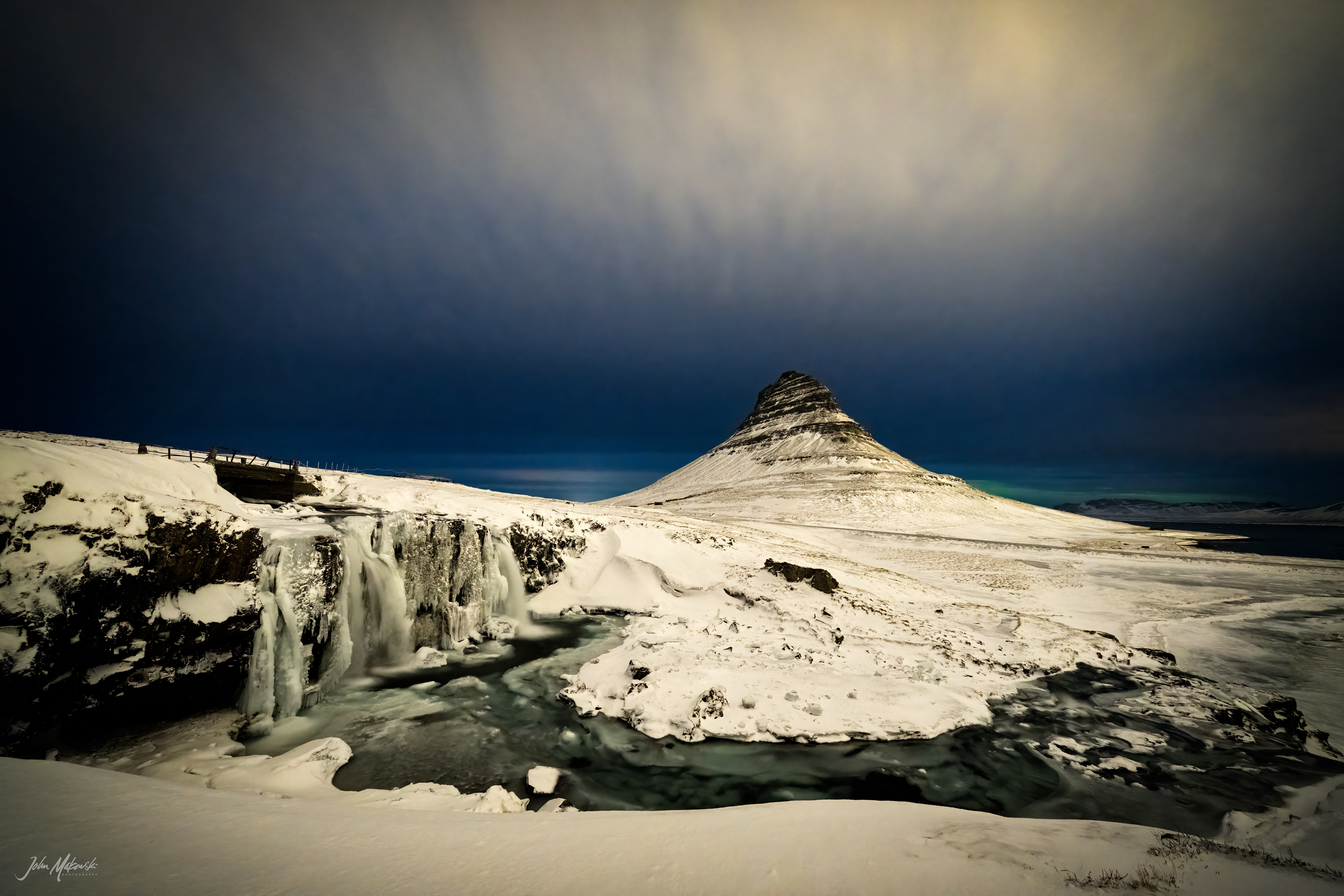 Kirkjufellsfoss at night with a sliver of Aurora Borealis on the horizon.