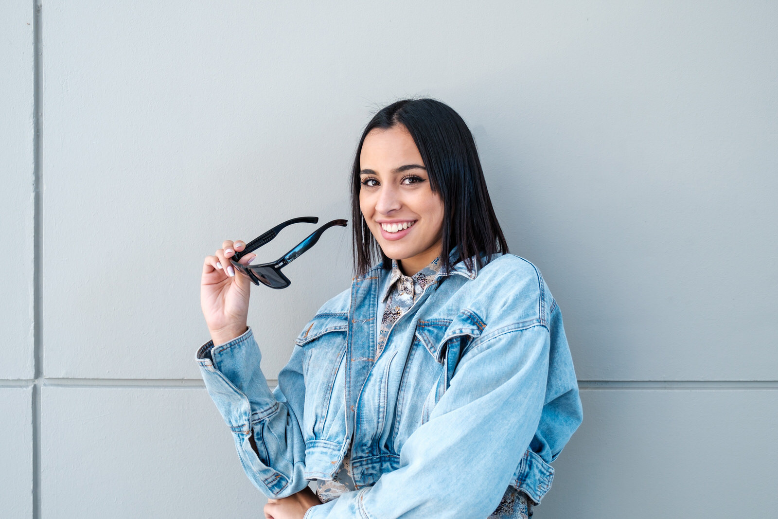 Smiling young woman holding sunglasses against a light gray wall, casual portrait