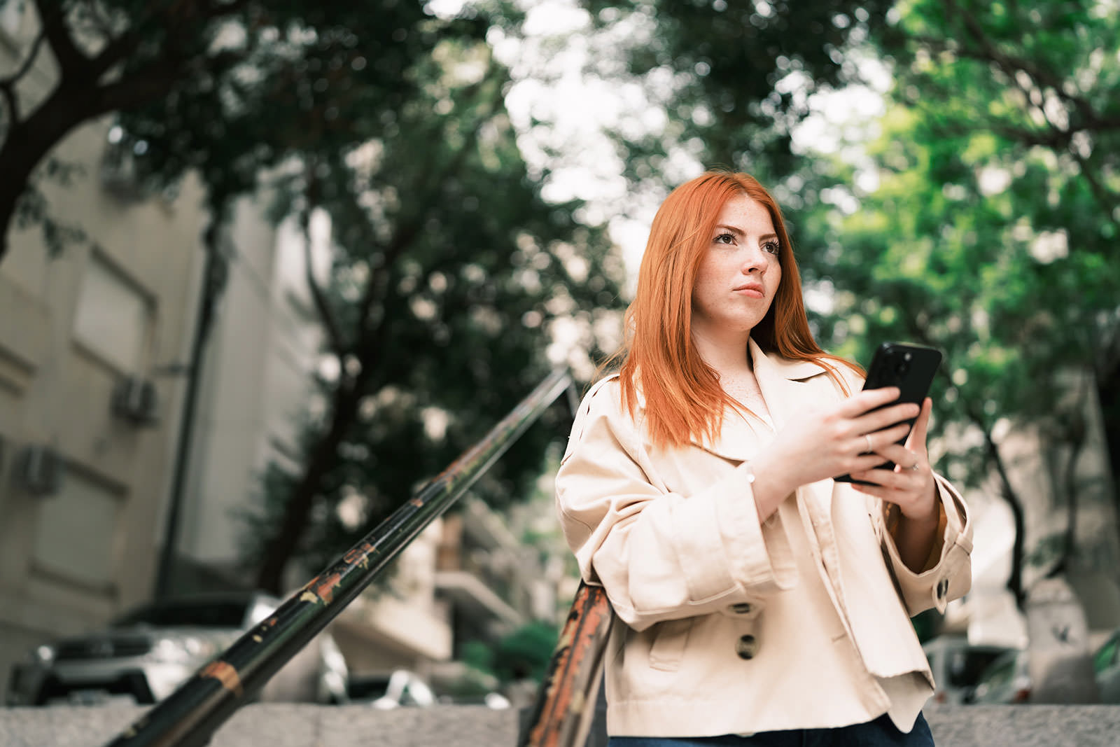 A red-haired woman holds a smartphone outdoors near a staircase, with trees and blurred buildings in the background