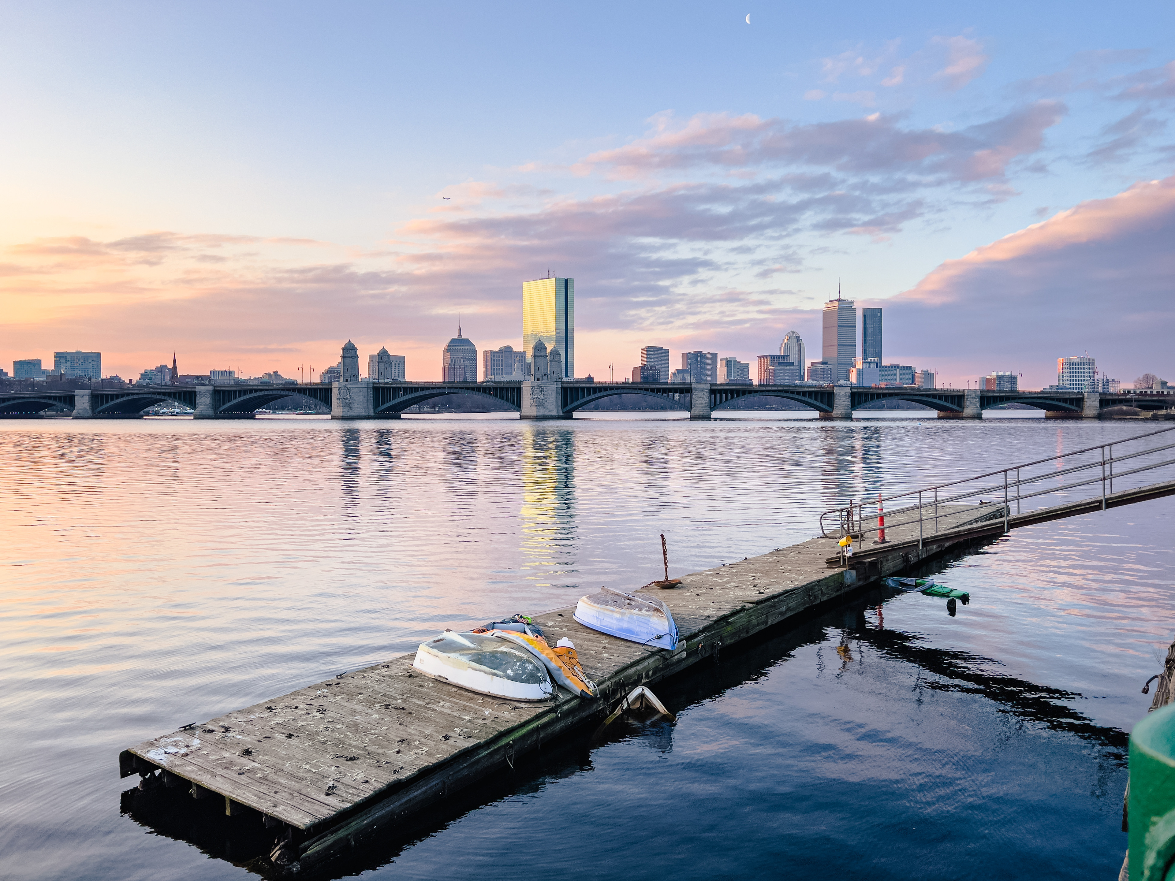 Sunrise illuminates Boston with the moon visible above a plane flying over a train seen over a boat on a dock in the Charles River.