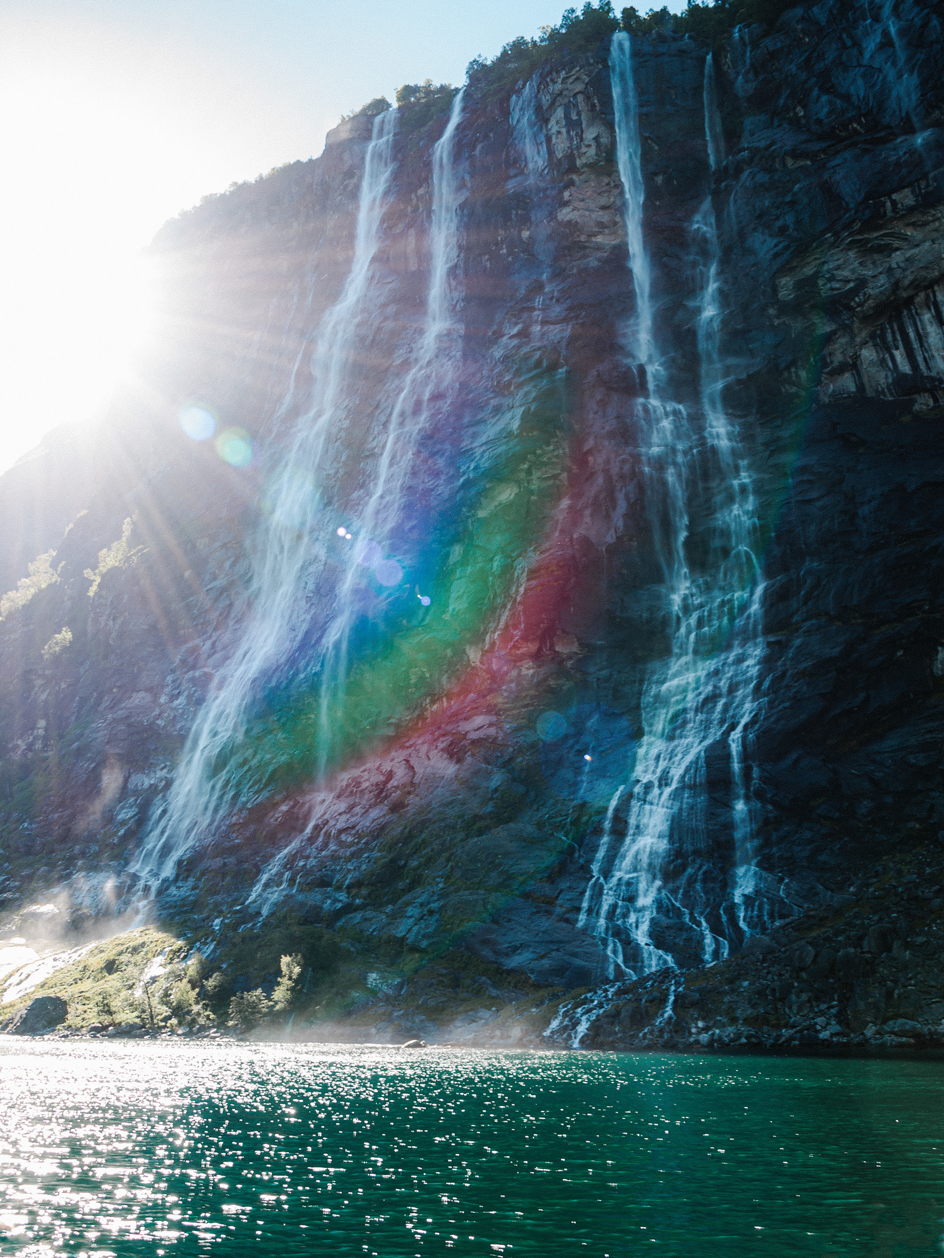 Sun rays burst through the mist to create a rainbow lens flare in Geirangerfjord