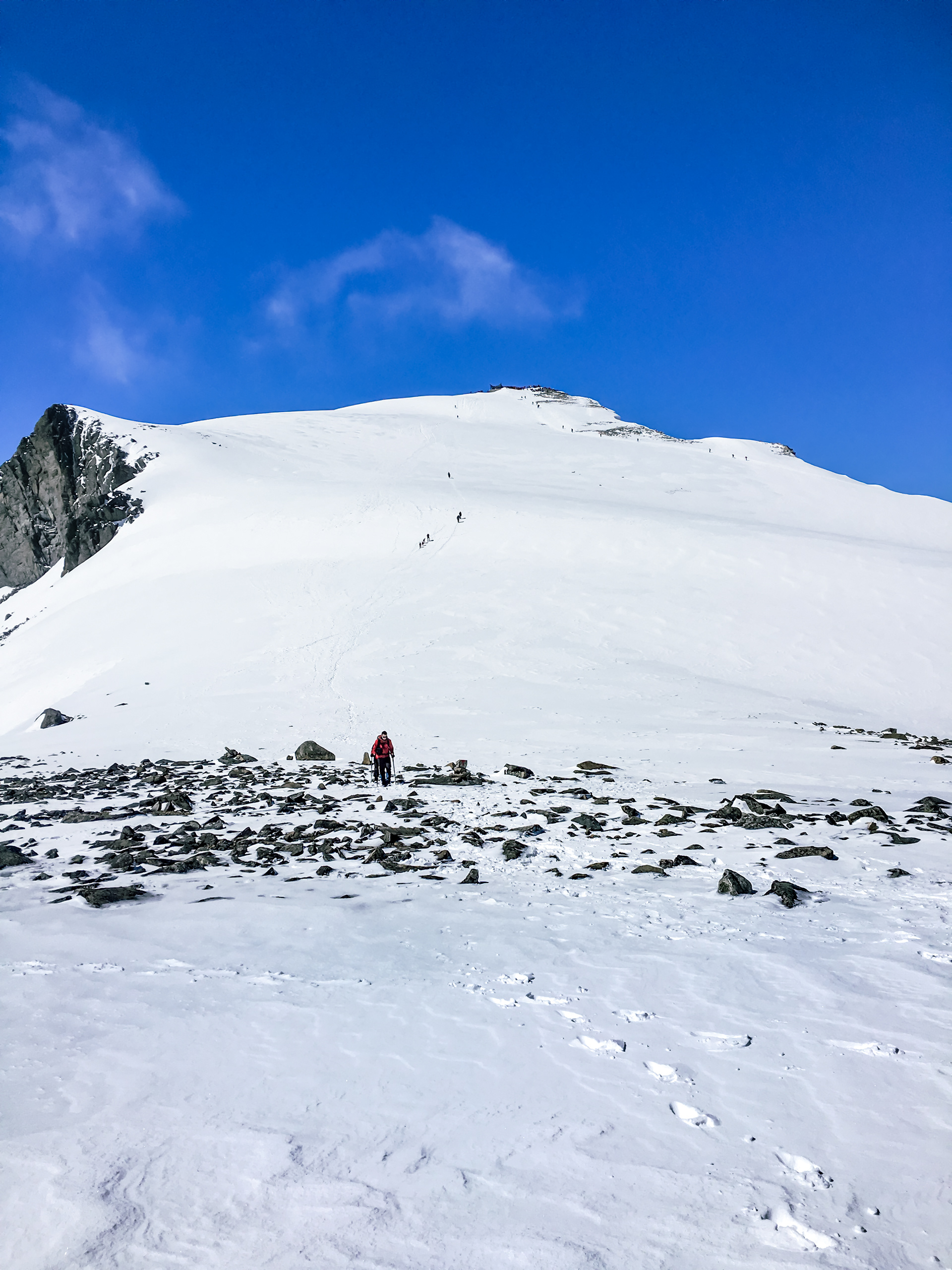 Two hikers starting their descent, with a distant view of hikers making their final push to the summit of Galdhøpiggen