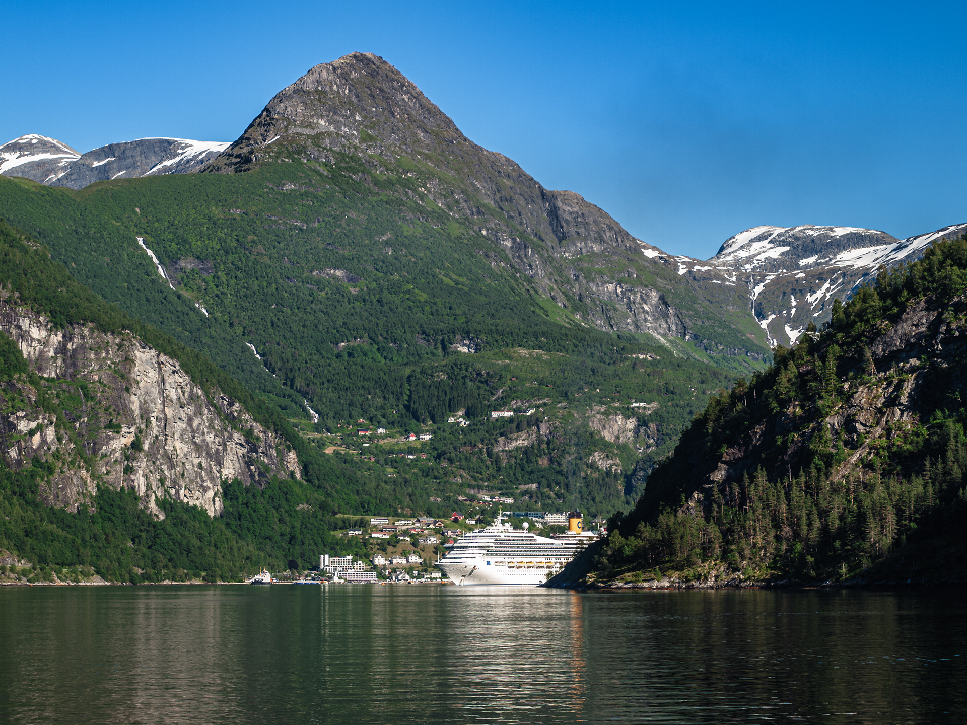 Large buildings in the port of Geirangerfjord are dwarfed by a giant cruise ship, itself dwarfed by a massive mountain towering behind