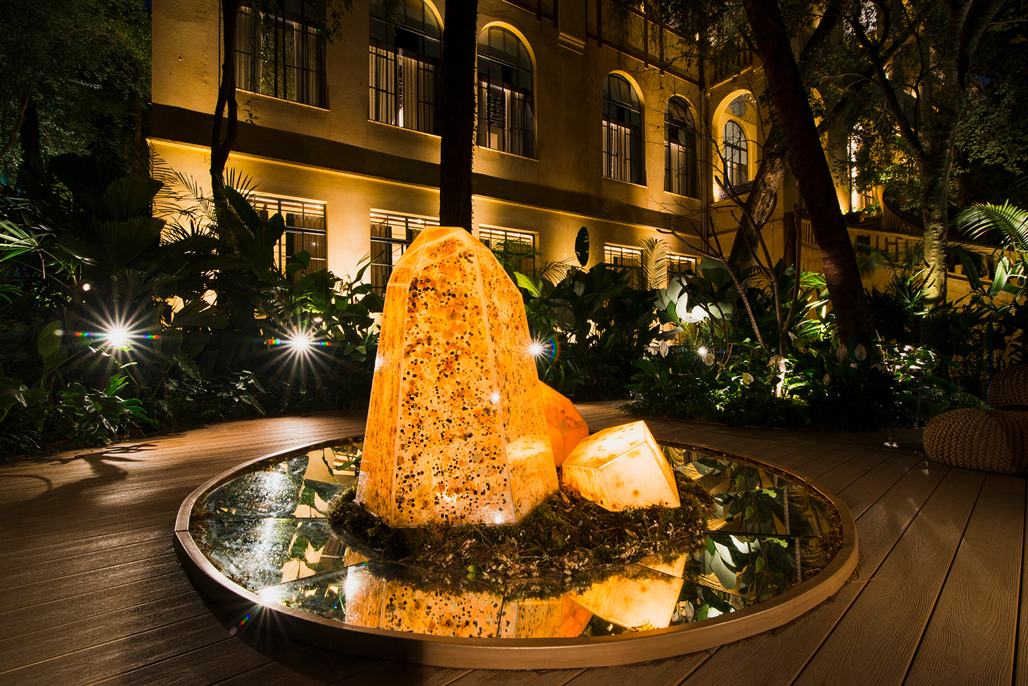 Instalação "O Avesso da Terra" de Bruna Mayer na CASACOR 2025, Parque Água Branca, São Paulo. Estrutura de cristais translúcidos sobre base circular espelhada, contendo microrganismos vivos cultivados, iluminada por refletores à noite, com vegetação ao redor. Installation "O Avesso da Terra" by Bruna Mayer at CASACOR 2025, Parque Água Branca, São Paulo. Structure of translucent crystals on a circular mirrored base, containing living cultivated microorganisms, illuminated by spotlights at night, surrounded by vegetation.