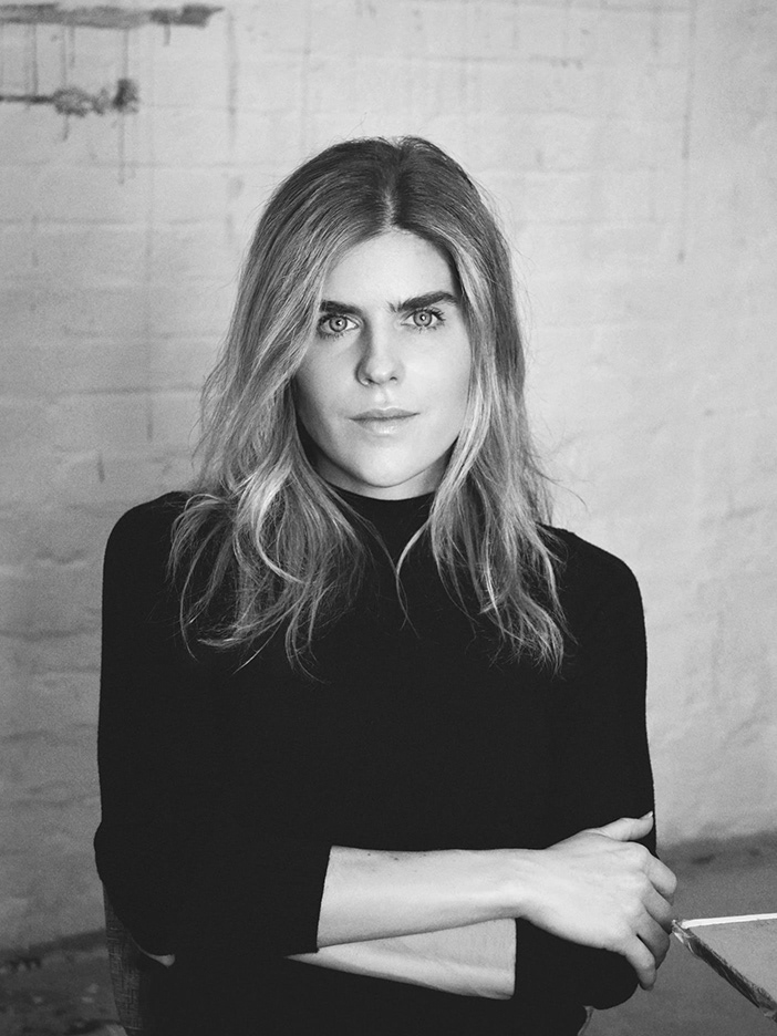 Retrato em preto e branco da artista visual Bruna Mayer, de pé, olhando para a câmera, com os braços cruzados, em frente a uma parede de tijolos. Black and white portrait of visual artist Bruna Mayer, standing with arms crossed, looking at the camera, in front of a brick wall.