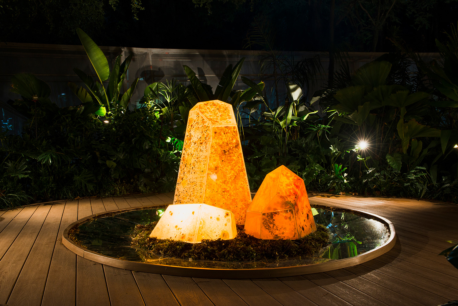 Instalação "O Avesso da Terra" de Bruna Mayer na CASACOR 2025, Parque Água Branca, São Paulo. Estrutura de cristais translúcidos sobre base circular espelhada, contendo microrganismos vivos cultivados, iluminada por refletores à noite, com vegetação ao redor. | Installation "O Avesso da Terra" by Bruna Mayer at CASACOR 2025, Parque Água Branca, São Paulo. Structure of translucent crystals on a circular mirrored base, containing living cultivated microorganisms, illuminated by spotlights at night, surrounded by vegetation.