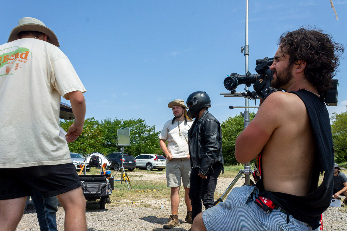 Ryder Sutton, Thomas Blanks, and a bearded Sam Casey with a farmer's tan and a muscle shirt, holding an Arri 16mm SRII film camera in front of an actor wearing motorcycle gear and a helmet on the set of "Forgive Me Father" with clear skies in Denton, Texas