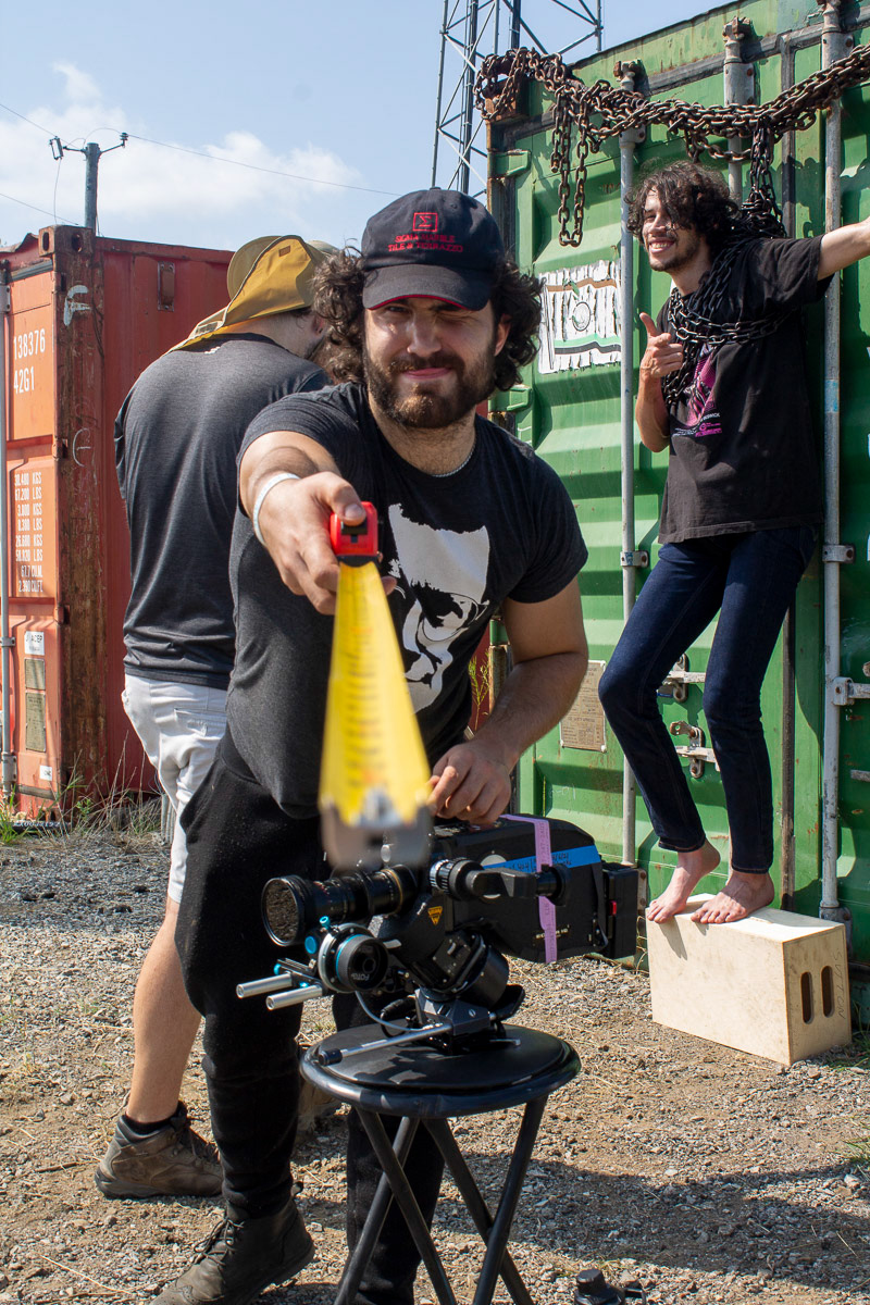 Sam Casey wearing a black-and-white shirt bearing an image of Del Close, standing above an Arri 16mm SRII film camera on a stool, he is holding measuring tape toward the photographer that is used to measure distance from the film plane indicator to the subject in focus, as an actor chained up to a storage container, standing on a full apple box standing on its side, on the set of "Forgive Me Father" on a partly cloudy North Texan day in Denton, Texas