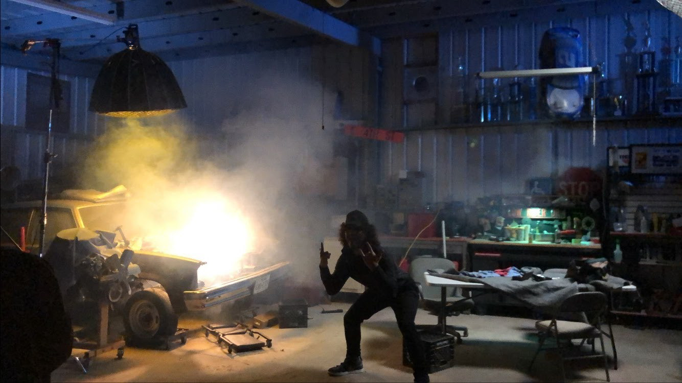 Sam Casey hunched in front of a fogged lighting setup in a car garage and holding up rock signs on the set of "All the Same"