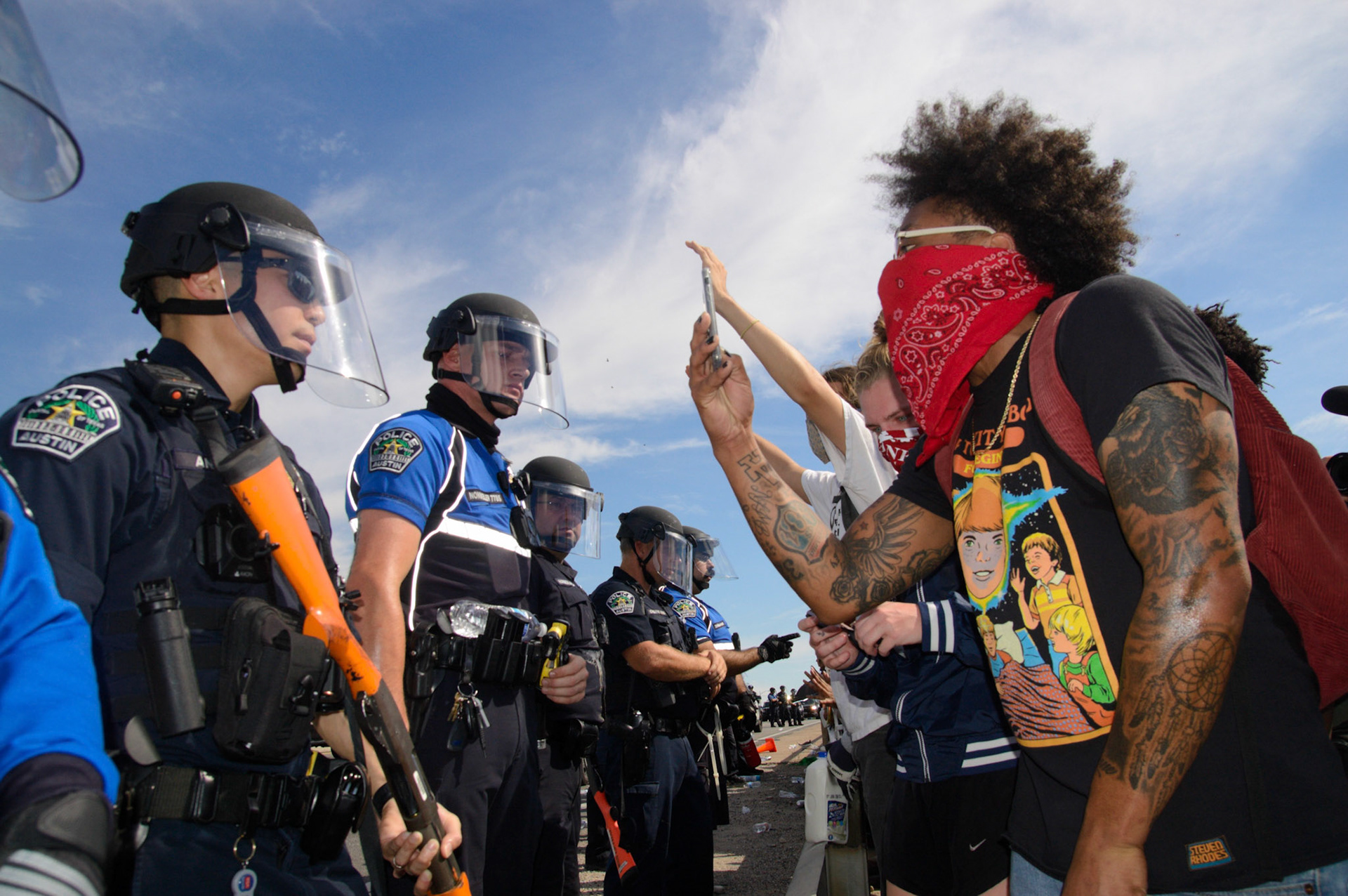 Photo by Sam Casey of protesters, one with a red bandana, filming police at a stopped intersection in Austin Texas after the Mike Ramos and George Floyd killings