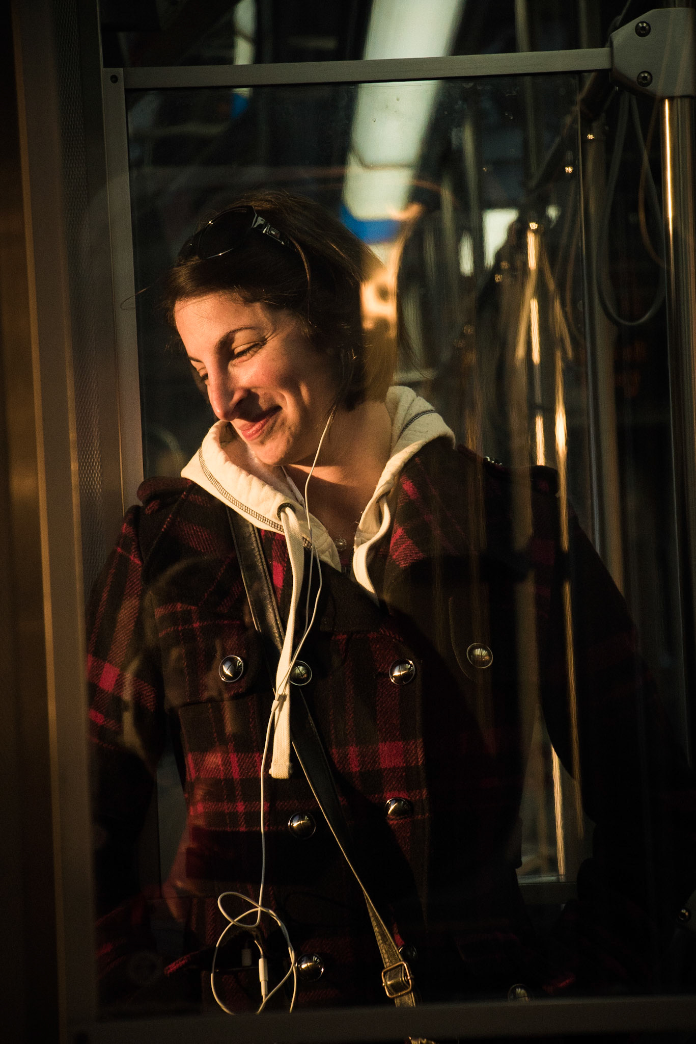 Photo of a woman through a pane of glass, smiling on a train wearing a red and black plaid wool coat over a white hoodie, with glasses in her hair during a phone call at sunset by Sam Casey