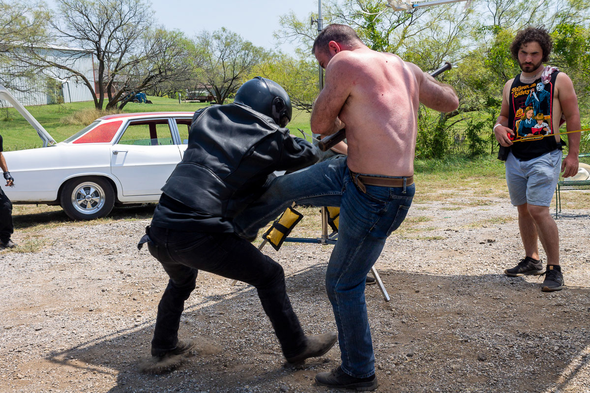 Casey holding tape measure for measuring distance from the film plane indicator to the subject while a shirtless man kicks a man dressed in black full motorcycle gear and helmet on the set of "Forgive Me Father" in Denton, Texas
