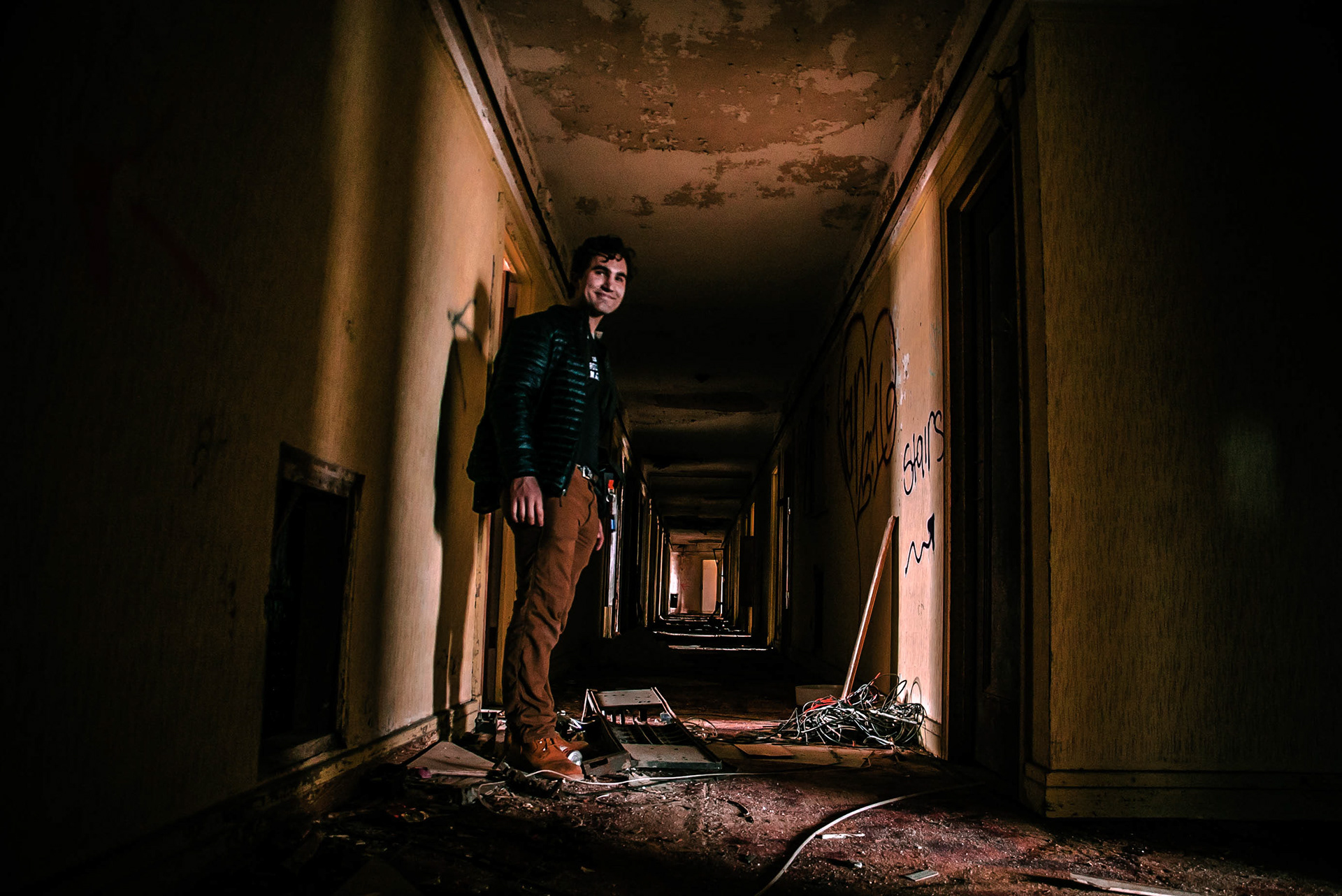 Sam Casey smiling in the middle of a derelict abandoned hotel hallway at sunset, filming for The Baker Hotel's renovation process in 2019 in Mineral Wells, Texas. The light casting becomes very contrasted in the hotel at times of dawn and dusk.