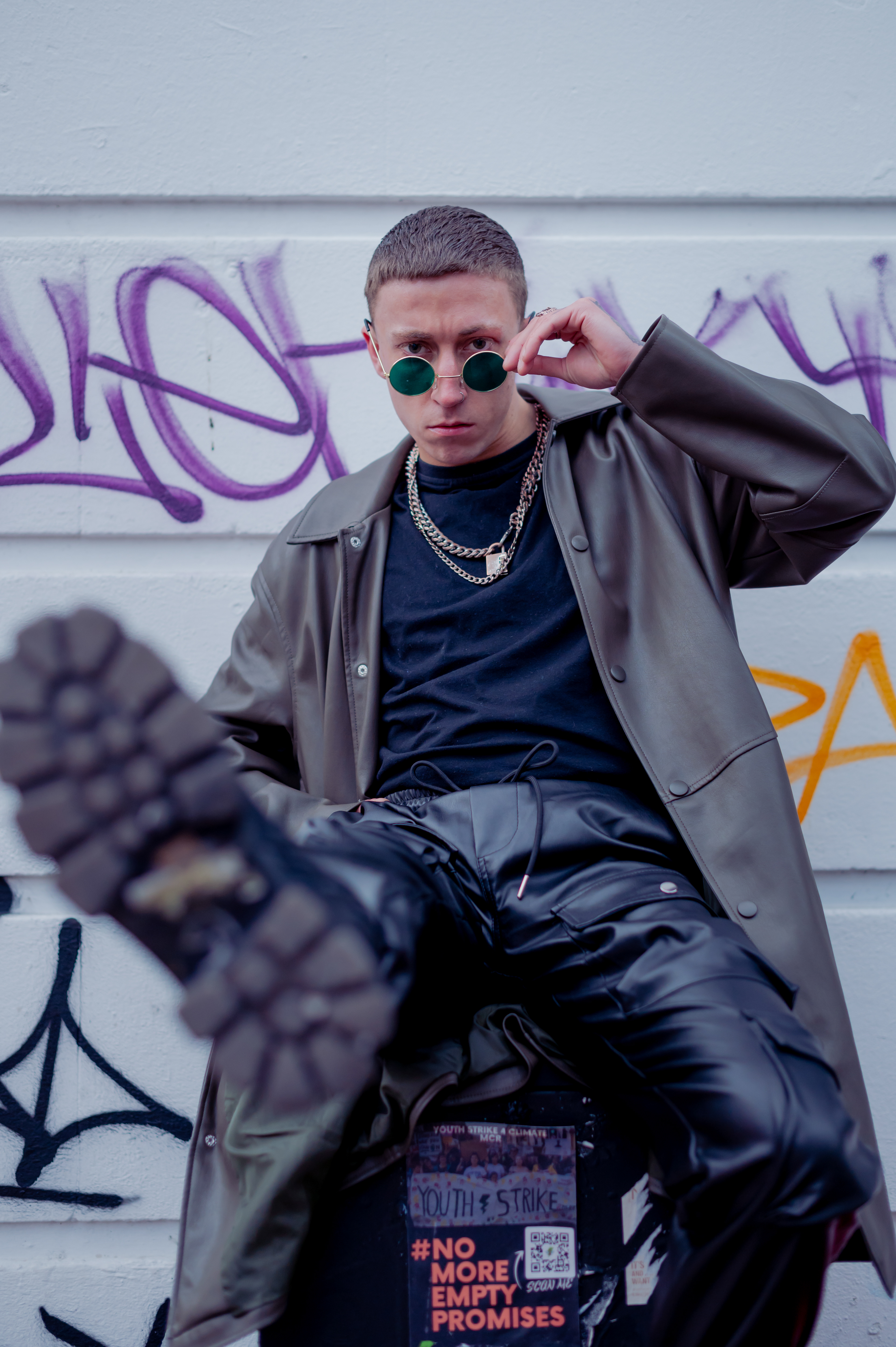 Man in boots, long leather jacket, leather pants, black t-shirt and chains, in Manchester - Northern Quarter , with sunglasses and rings 