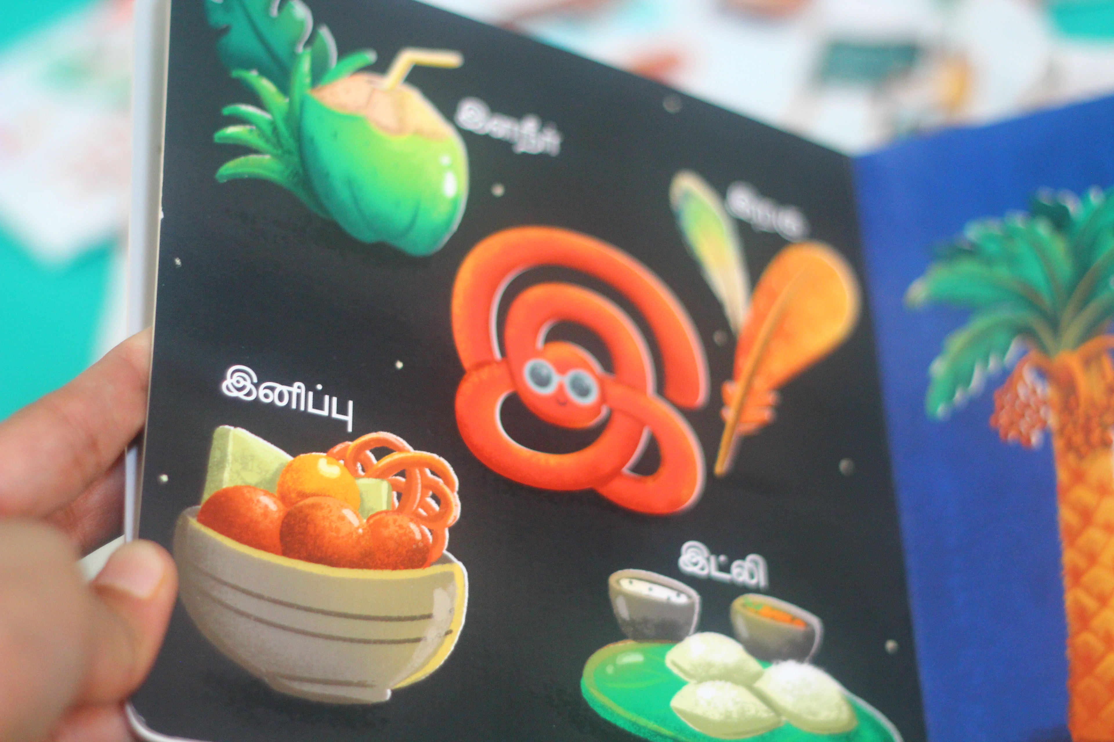 Uyirum Tamizhum Tamil vowels board book: child's hand on a page with fruit and sweets