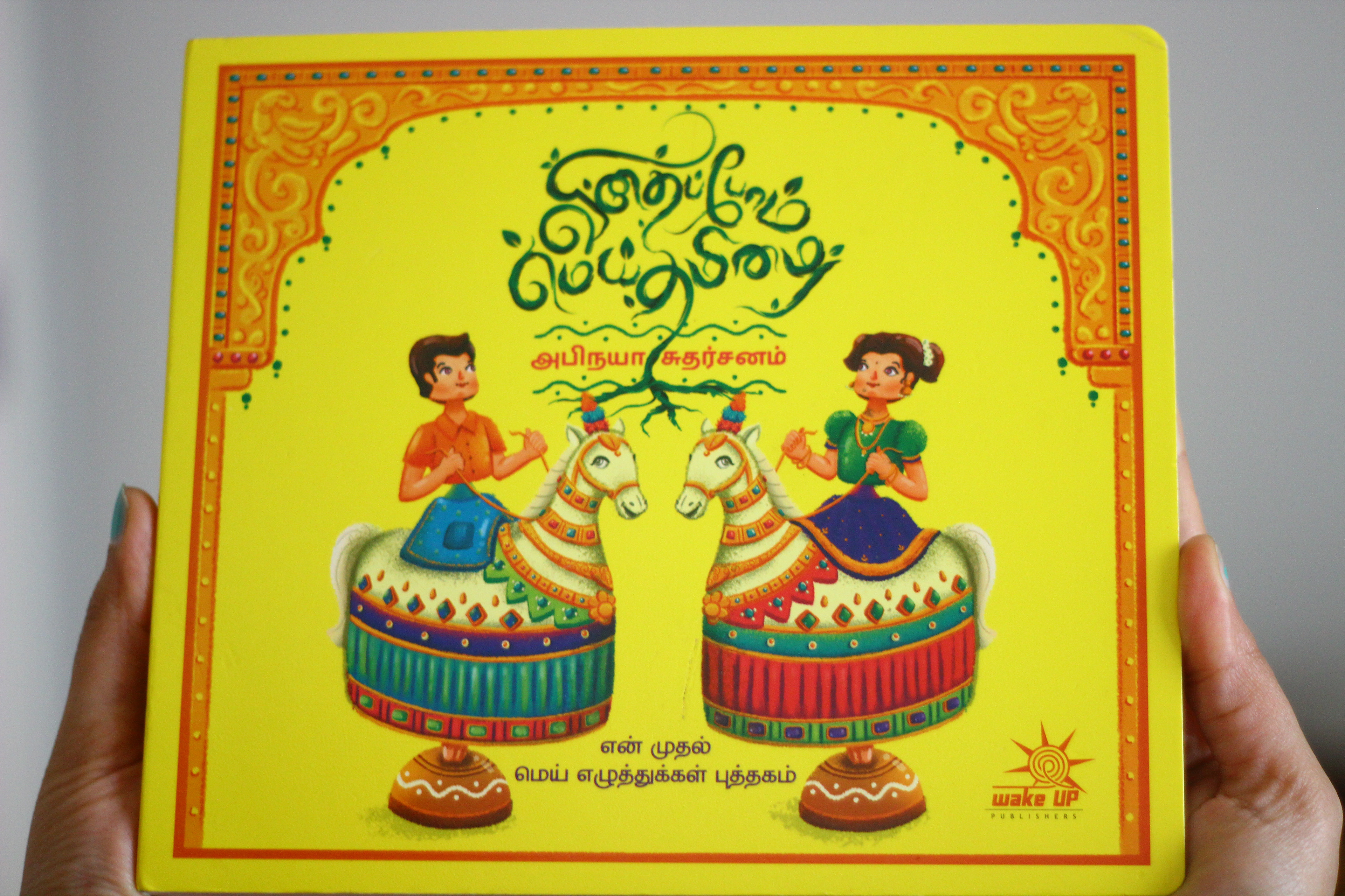 Vidhaippom Meithamizhai Tamil alphabet board book: hands holding the yellow board book cover, illustration by Atma Studios