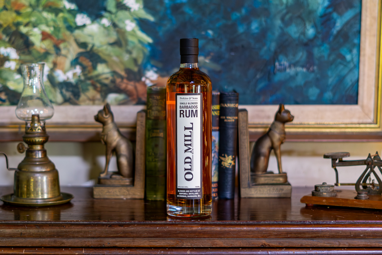 Old Mill Barbados Rum bottle displayed on an antique wooden shelf with vintage ornaments and books