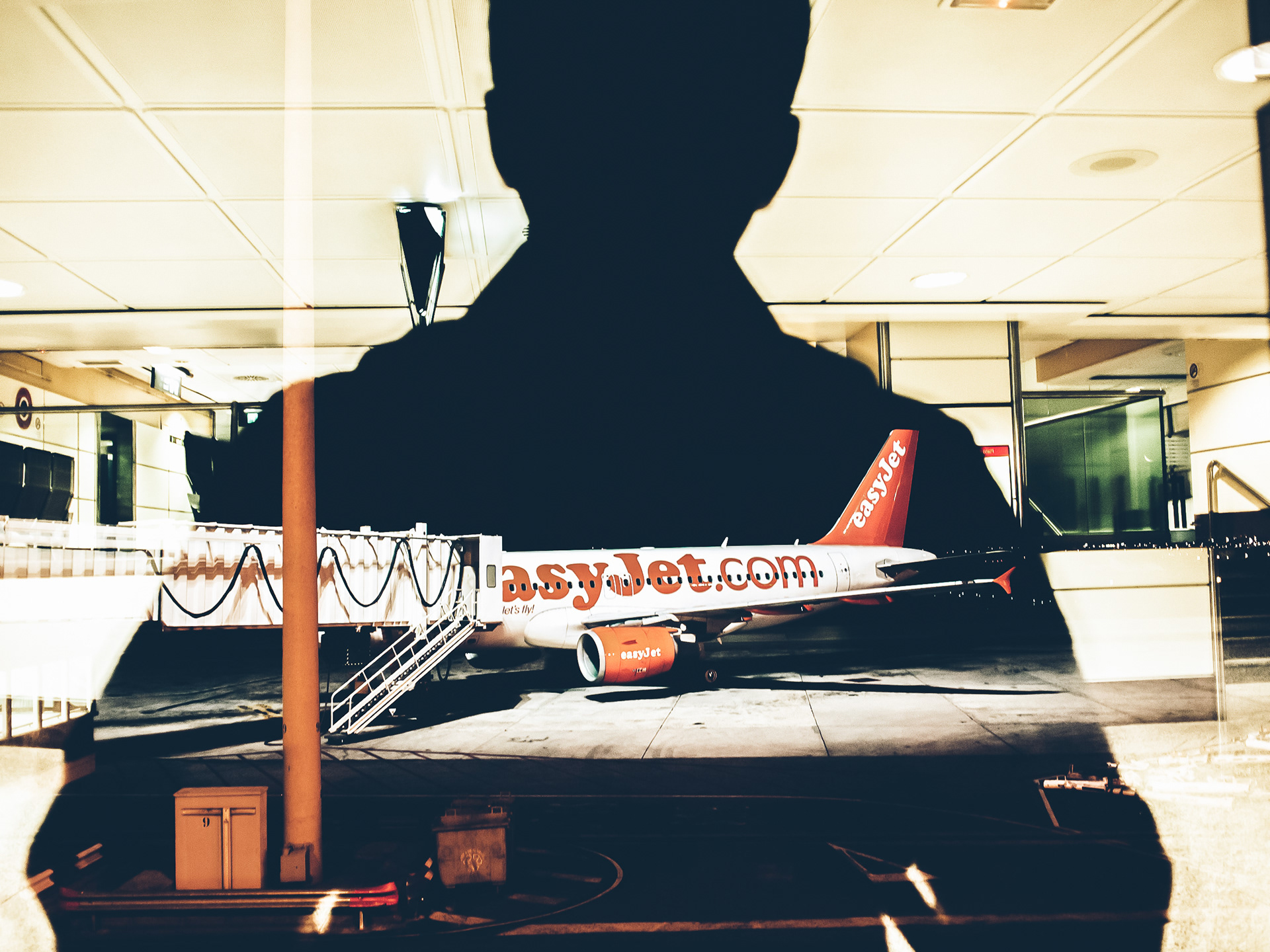 Mahesh Shantaram seen reflected as a silhoutte in the window of an airport terminal overlooking an EasyJet flight waiting on the tarmac.