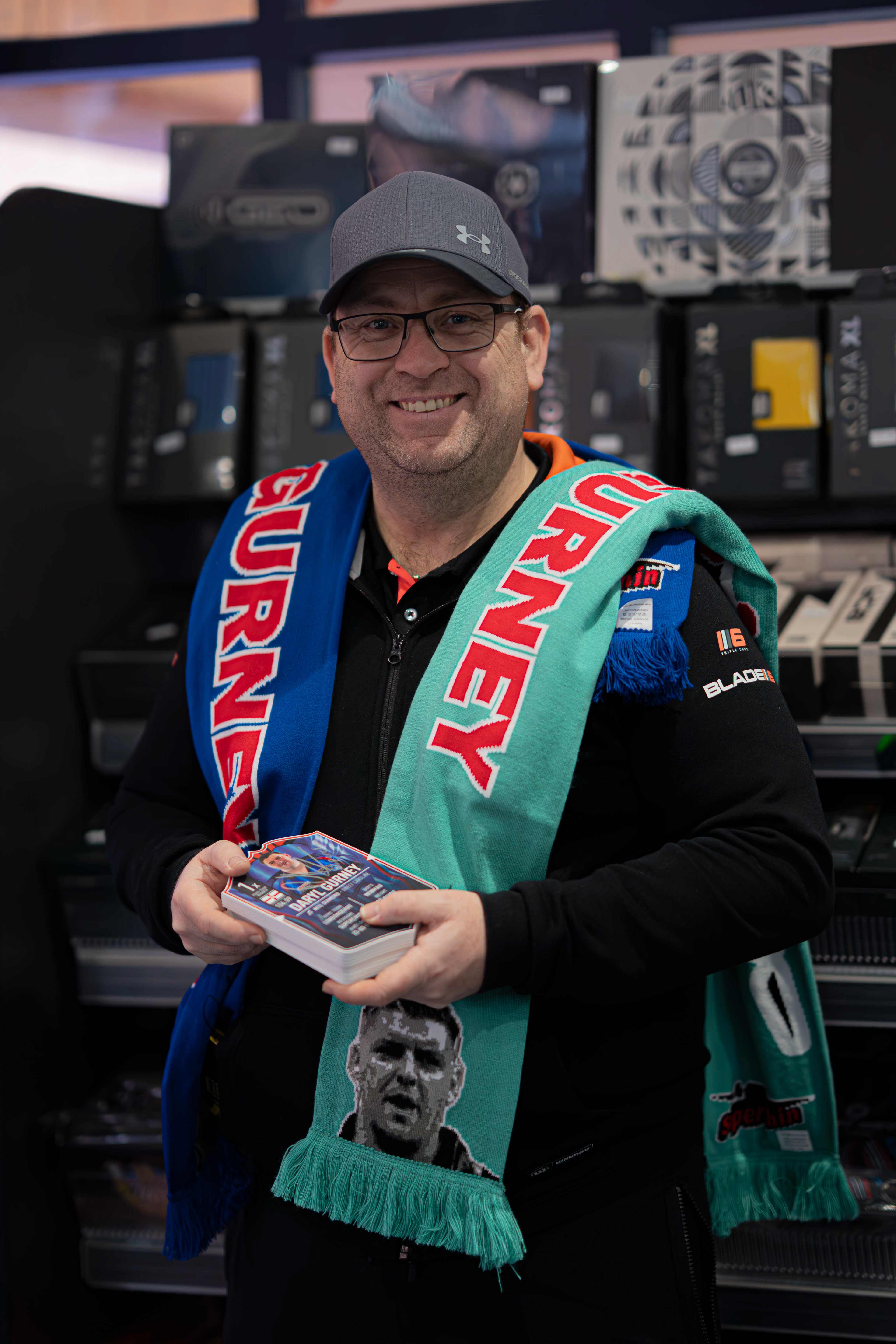 Staff member standing next to Daryl Gurney holding merch for fans to buy at Midlands Darts Shop, Tamworth, Staffordshire. 1st March 2026