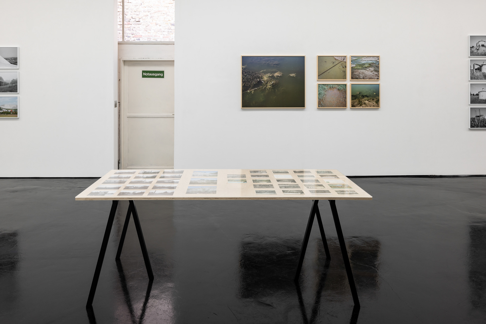 Raumansicht mit Tisch auf dem Found Footage Postkarten ausgestellt sind und gerahmten Fotografien an der Wand zu Gewässerbelastung