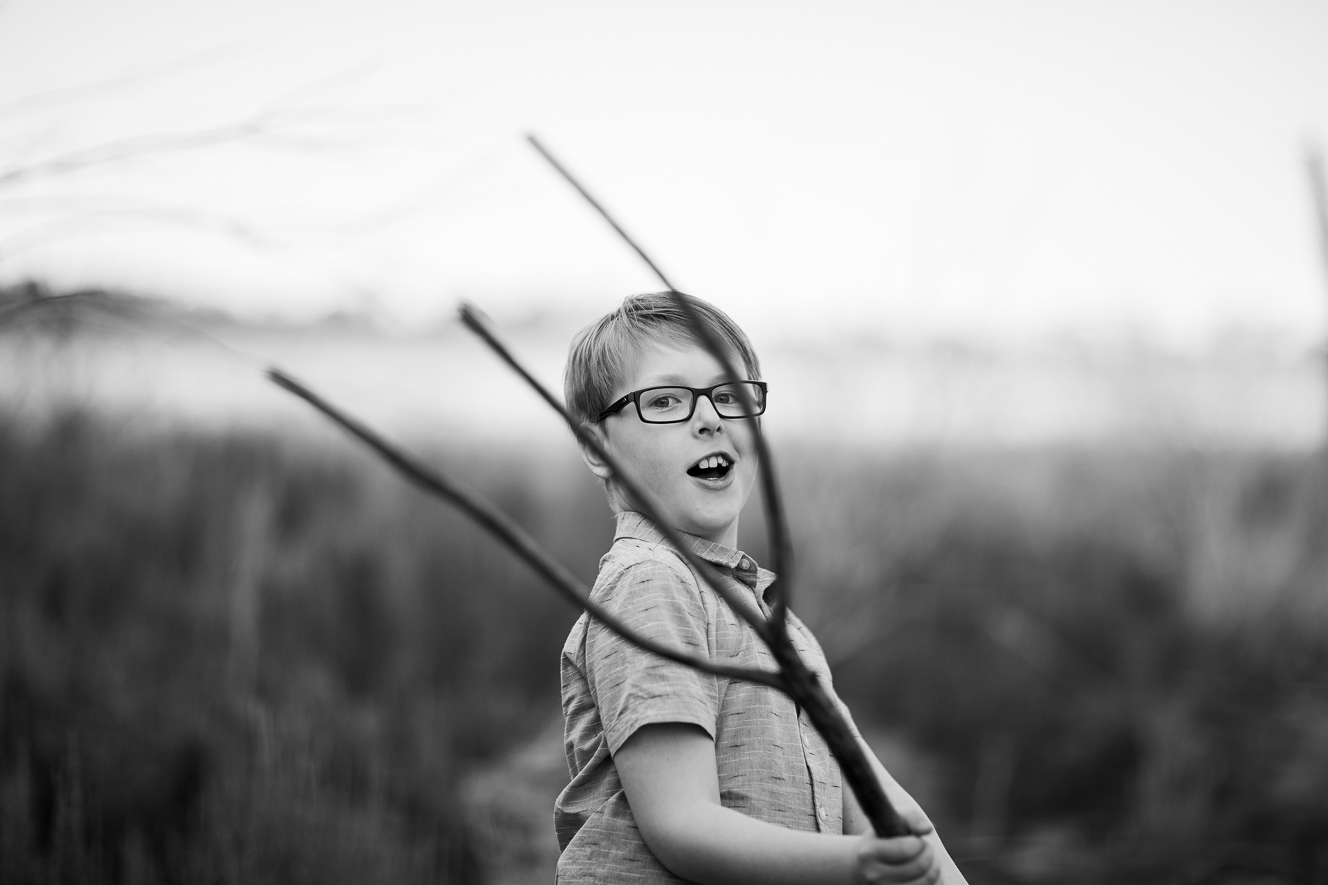 portrait photo of a boy in a shirt with water and tree background. looking through a fork shaped stick in a playful manner. Black and white image 