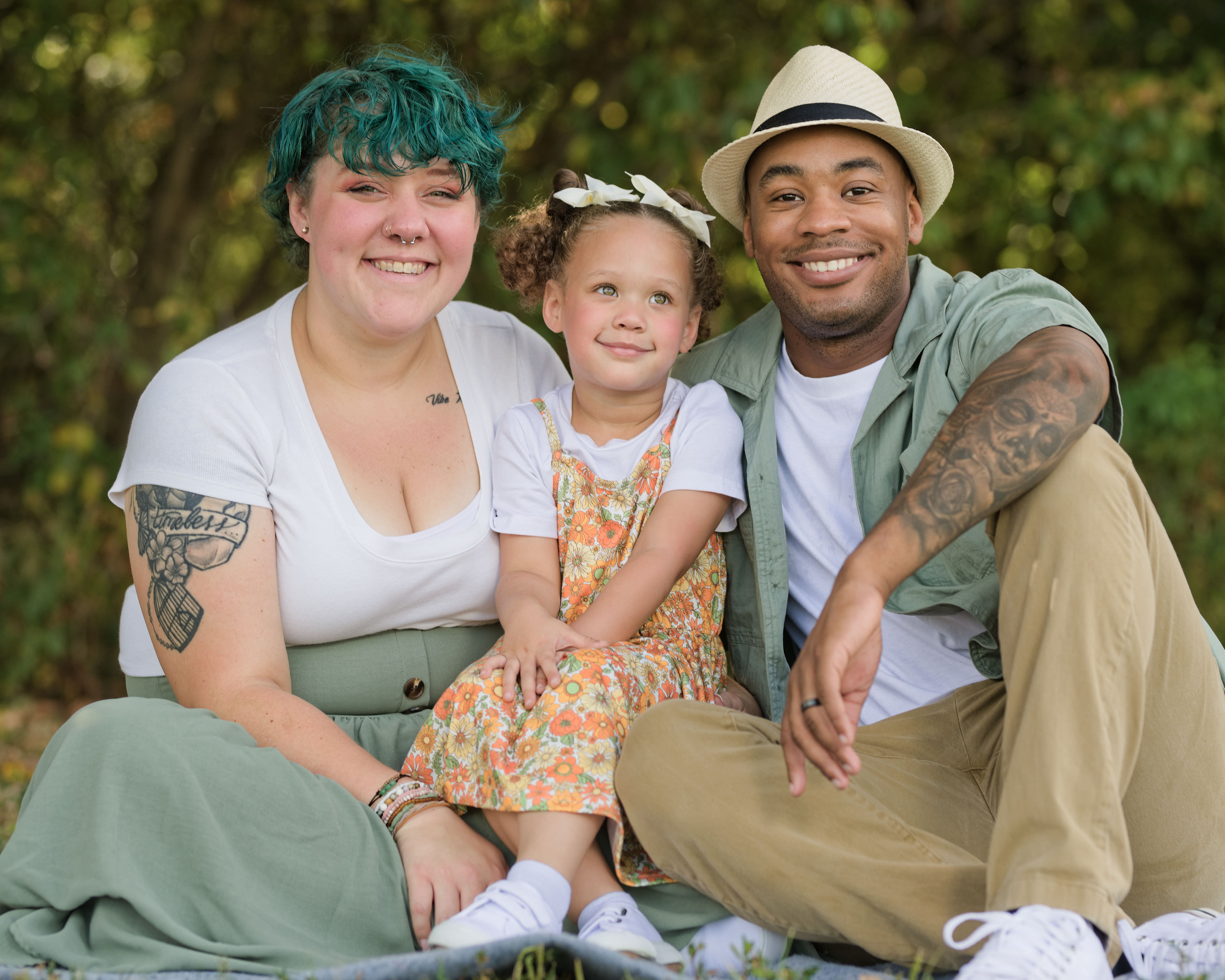 Man, woman, and young girl seated on a blanket outdoors in a green park setting—the man and woman both have visible tattoos and casual attire, while the girl wears a floral dress over a white shirt. Captured in soft natural light, conveying warmth, individuality, and togetherness.