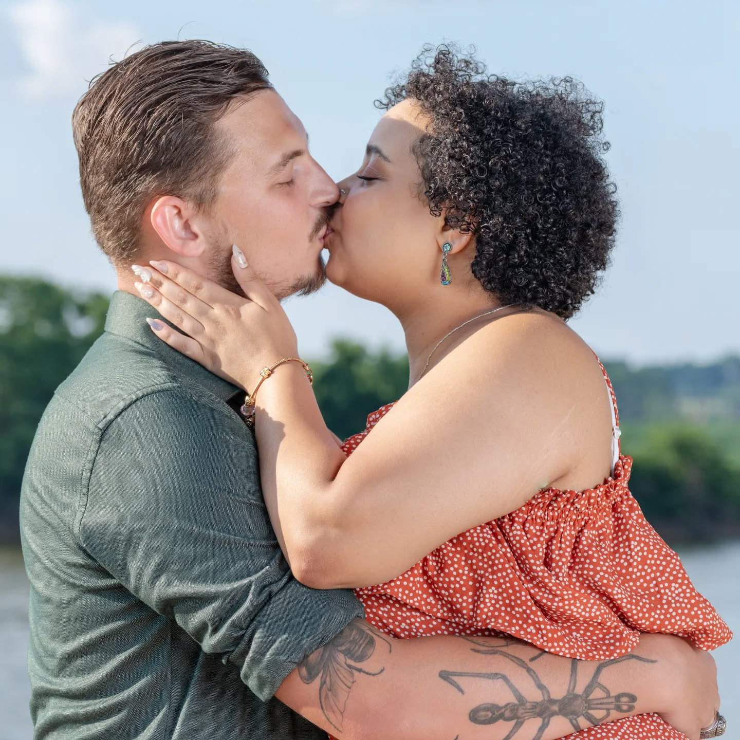 Man and woman sharing a posed kiss by a wooded lake—she wears a red polka dot dress and cradles his face, while he stands in a green shirt with tattooed arms. Surrounded by trees and calm water, the moment feels romantic, grounded, and serene.