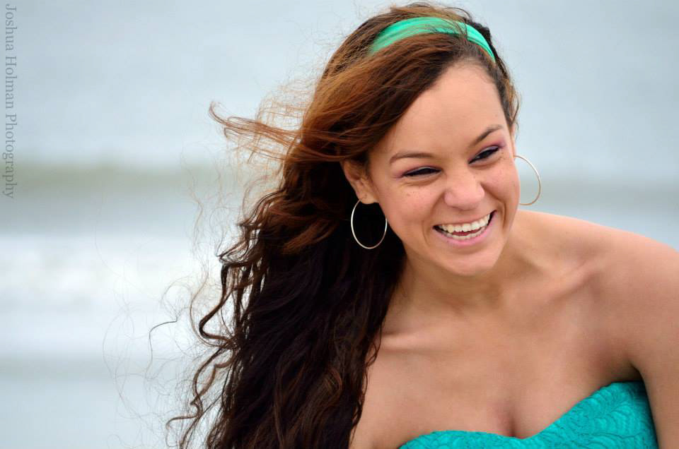 Senior portrait of a girl on a beach with ocean waves in the background, featuring a person with long wavy hair, hoop earrings, and a strapless turquoise top—natural light and confident gaze create a relaxed, summery mood.
