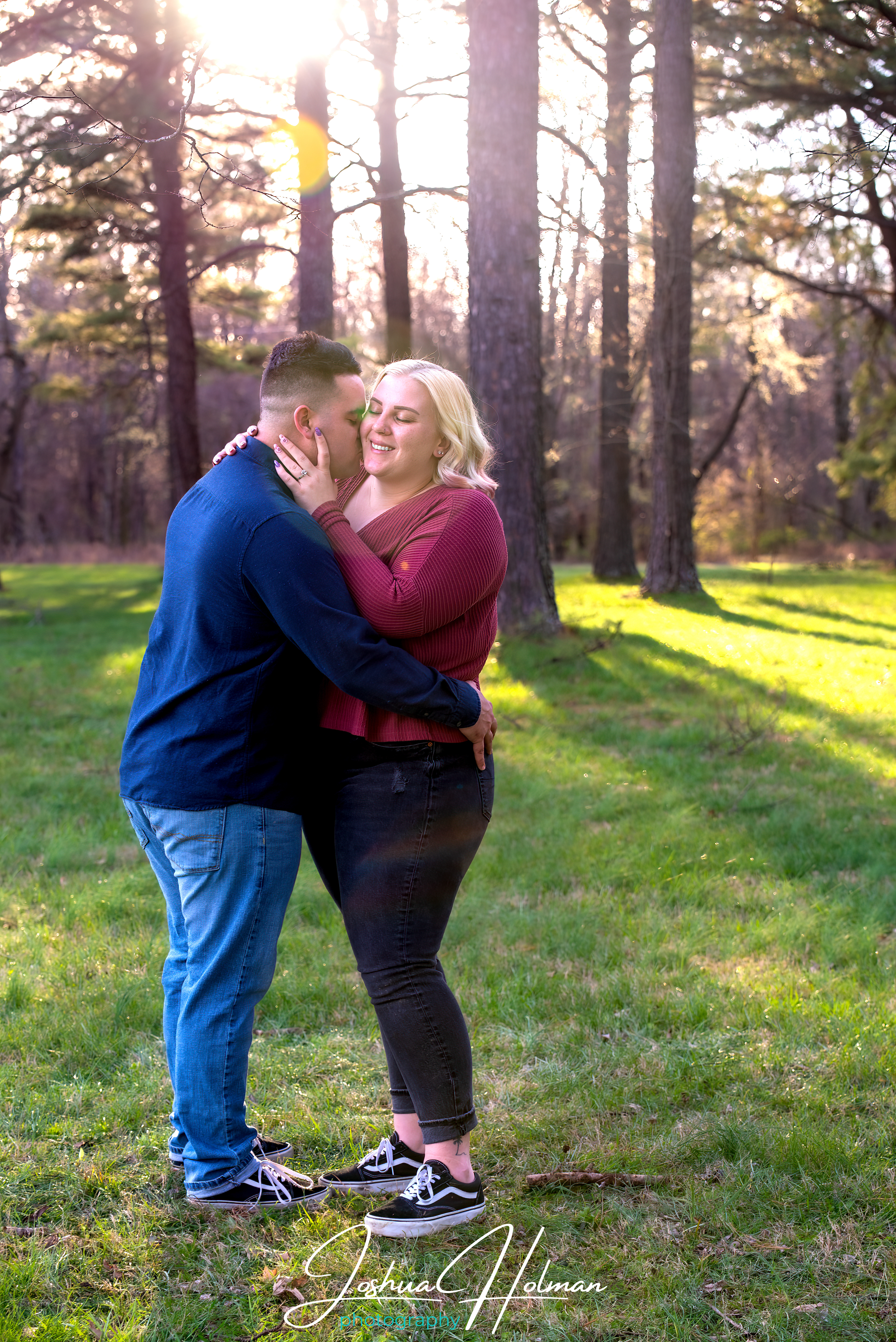 Man and woman standing close together in a sunlit forest clearing—embracing with heads gently touching, surrounded by tall trees and soft afternoon light, capturing tenderness and quiet connection.