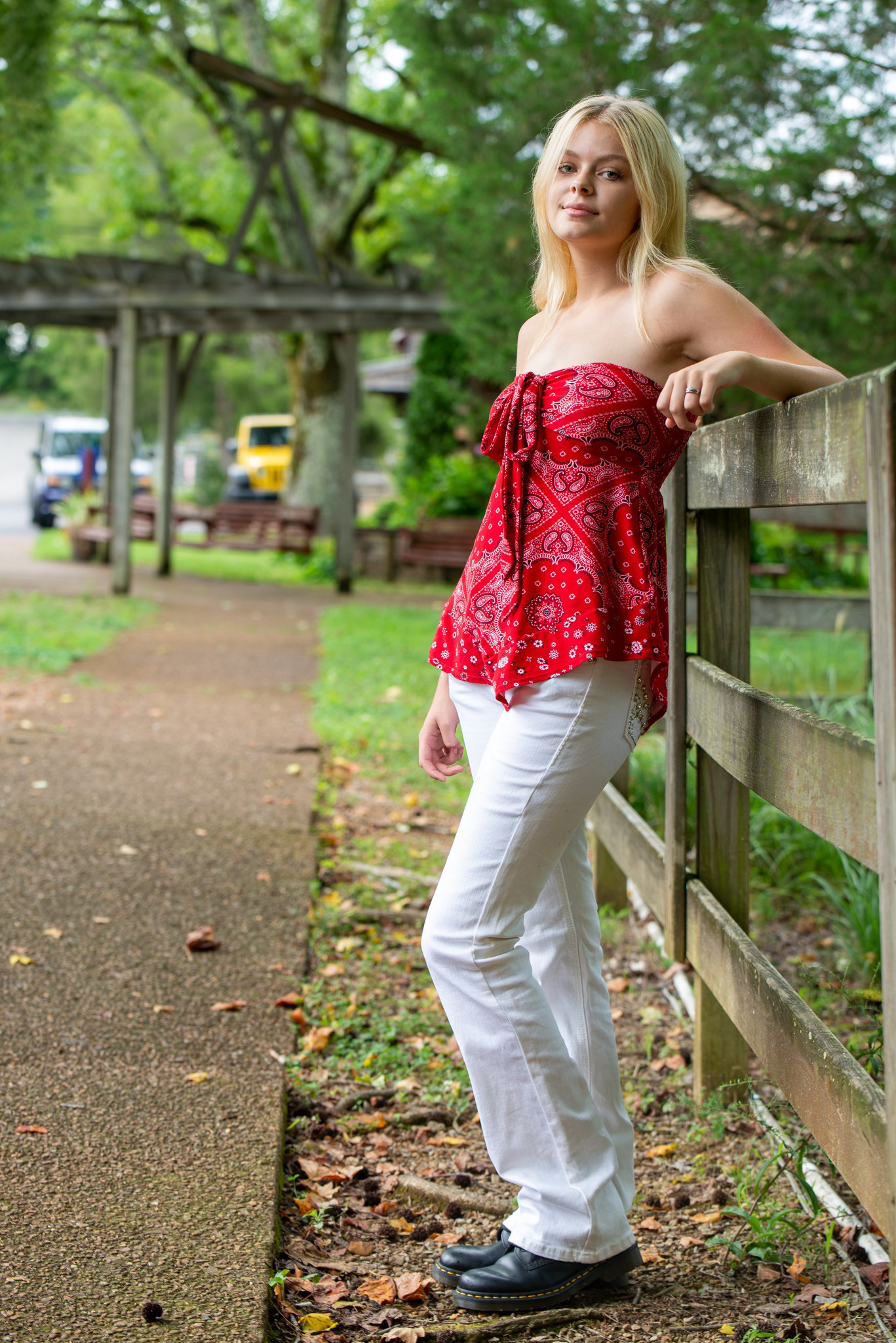 Senior portrait of a girl outdoors with warm, golden light—subject leaning against a wooden fence, wearing a red strapless top with a bow and white pants, framed by a sunlit path and soft greenery.
