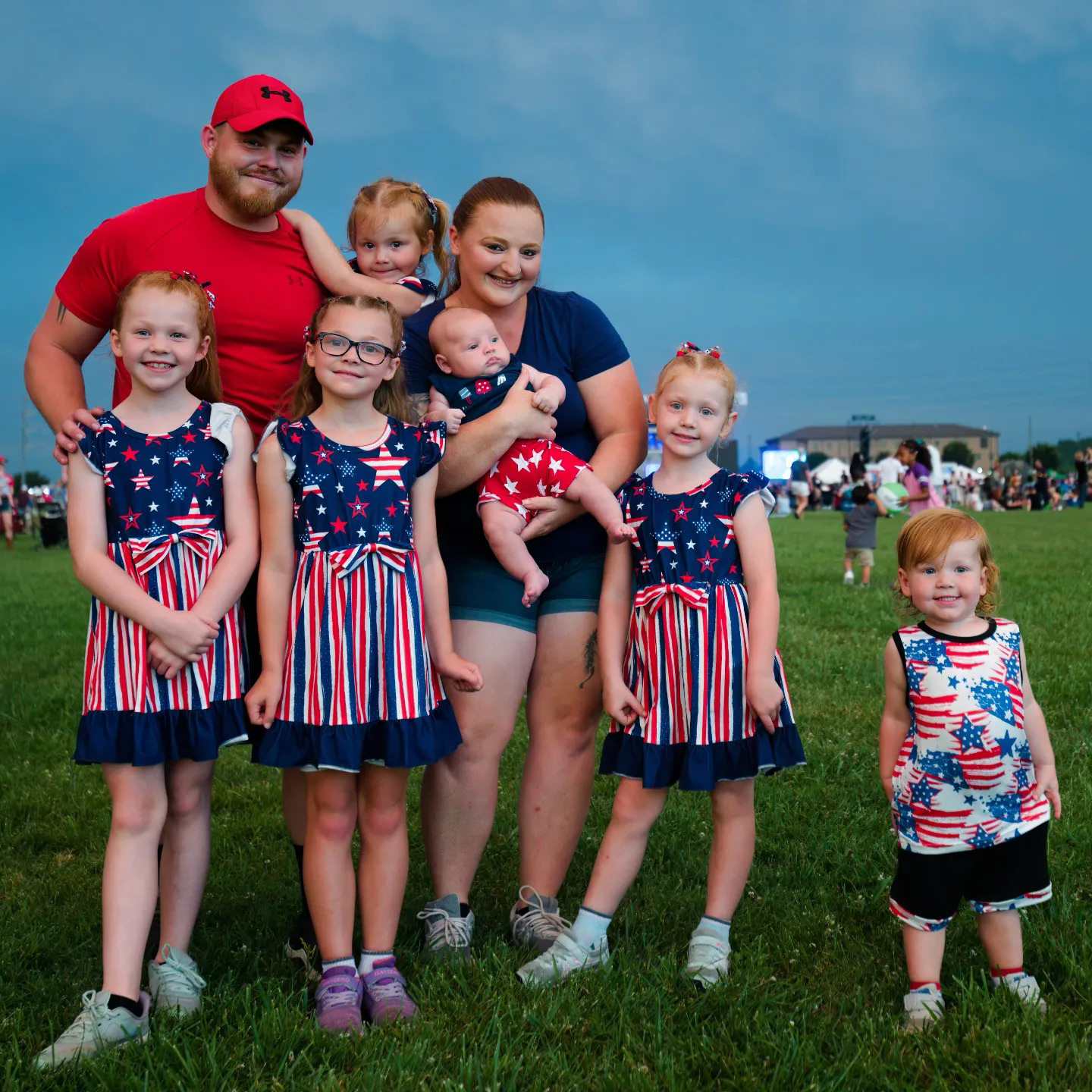 Family of seven standing together in a grassy park—two parents and five children, with the kids dressed in matching patriotic outfits featuring stars and stripes. Captured during a festive outdoor gathering, radiating joy, unity, and belonging.