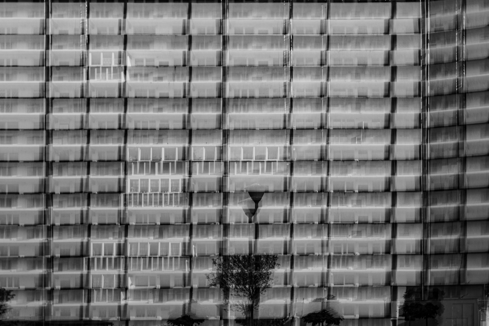 Black-and-white abstract photograph of a modern high-rise apartment building facade with repeating balconies, motion blur, and layered reflections creating a geometric architectural pattern.