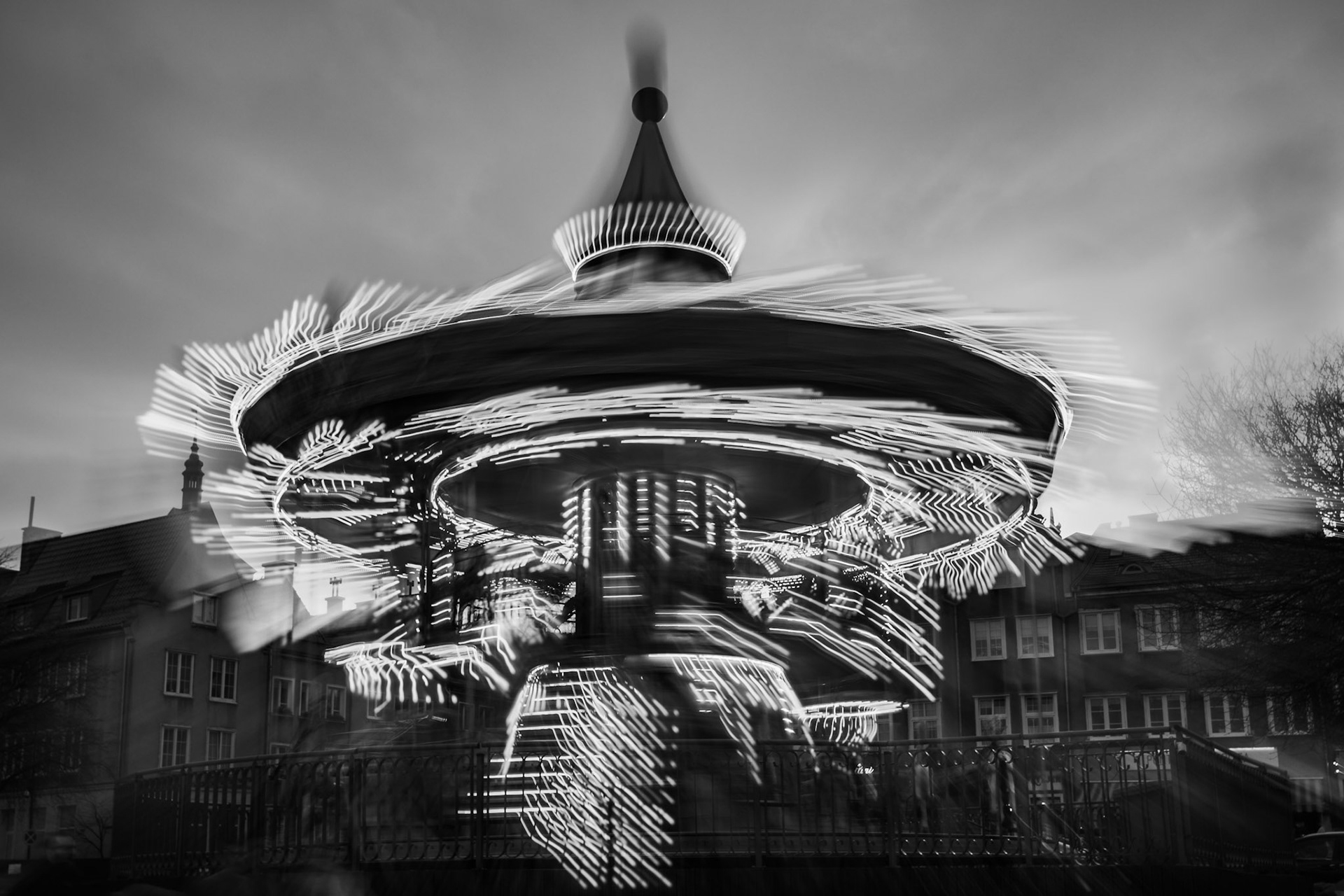 Treść wiadomości Gemini A long exposure black and white photograph of a spinning carousel in a European city square, featuring dramatic motion blur and glowing light trails against a backdrop of historic buildings.