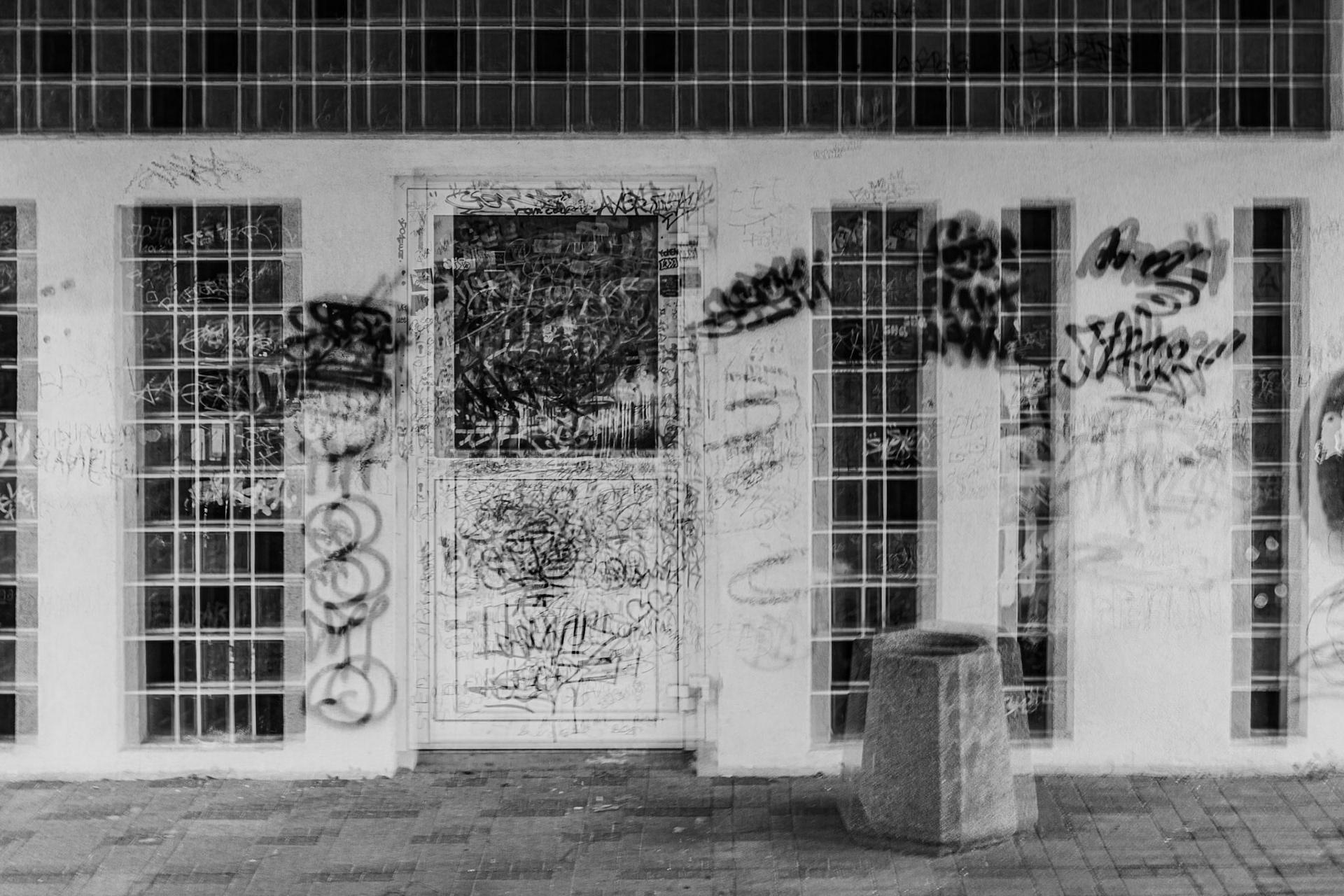 Black-and-white photo of a graffiti-covered urban building entrance with tall grid windows, tagged walls, and a concrete trash bin on a city sidewalk.
