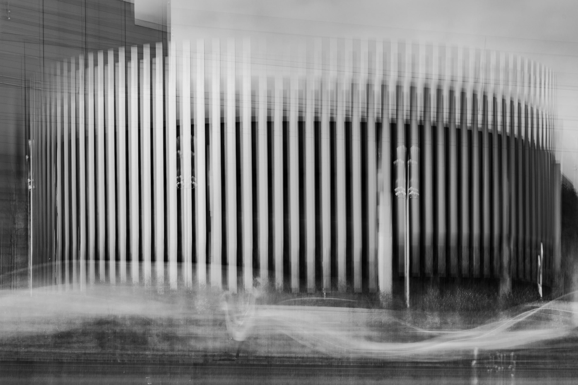 Abstract black-and-white motion blur photo of a modern building with vertical columns and ghosted figures, creating a dynamic architectural scene.