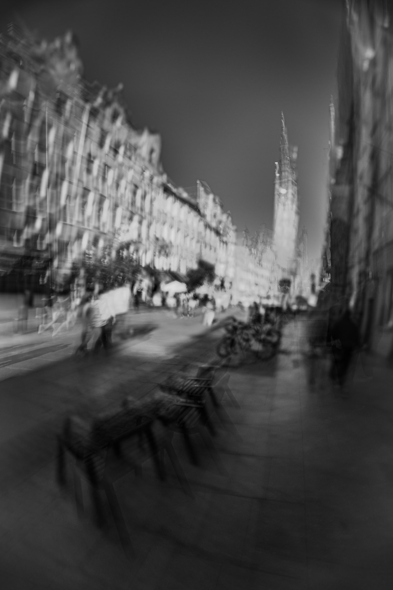 Black-and-white long-exposure street scene with motion blur, showing historic city buildings, pedestrians, outdoor seating, and a cathedral spire in the distance, creating a dreamlike urban atmosphere.