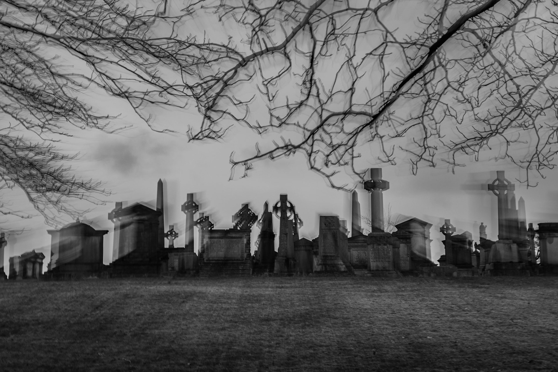 Blurry black and white long exposure of Celtic crosses and graves in a cemetery under bare, skeletal tree branches.