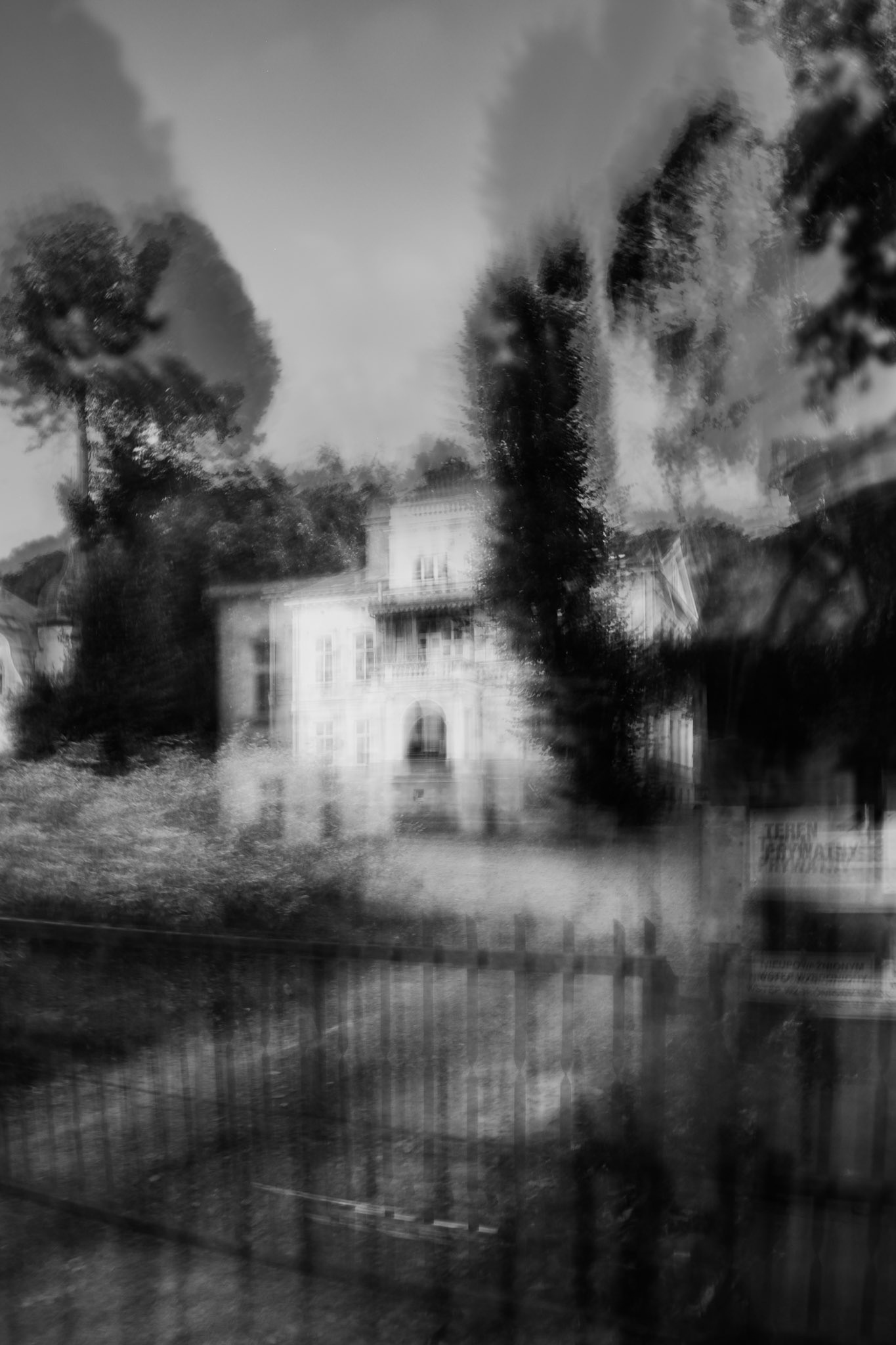 Black and white blurred photo of an old house surrounded by trees, with a fence in the foreground, creating a mysterious and haunting atmosphere, made in ICM technique.