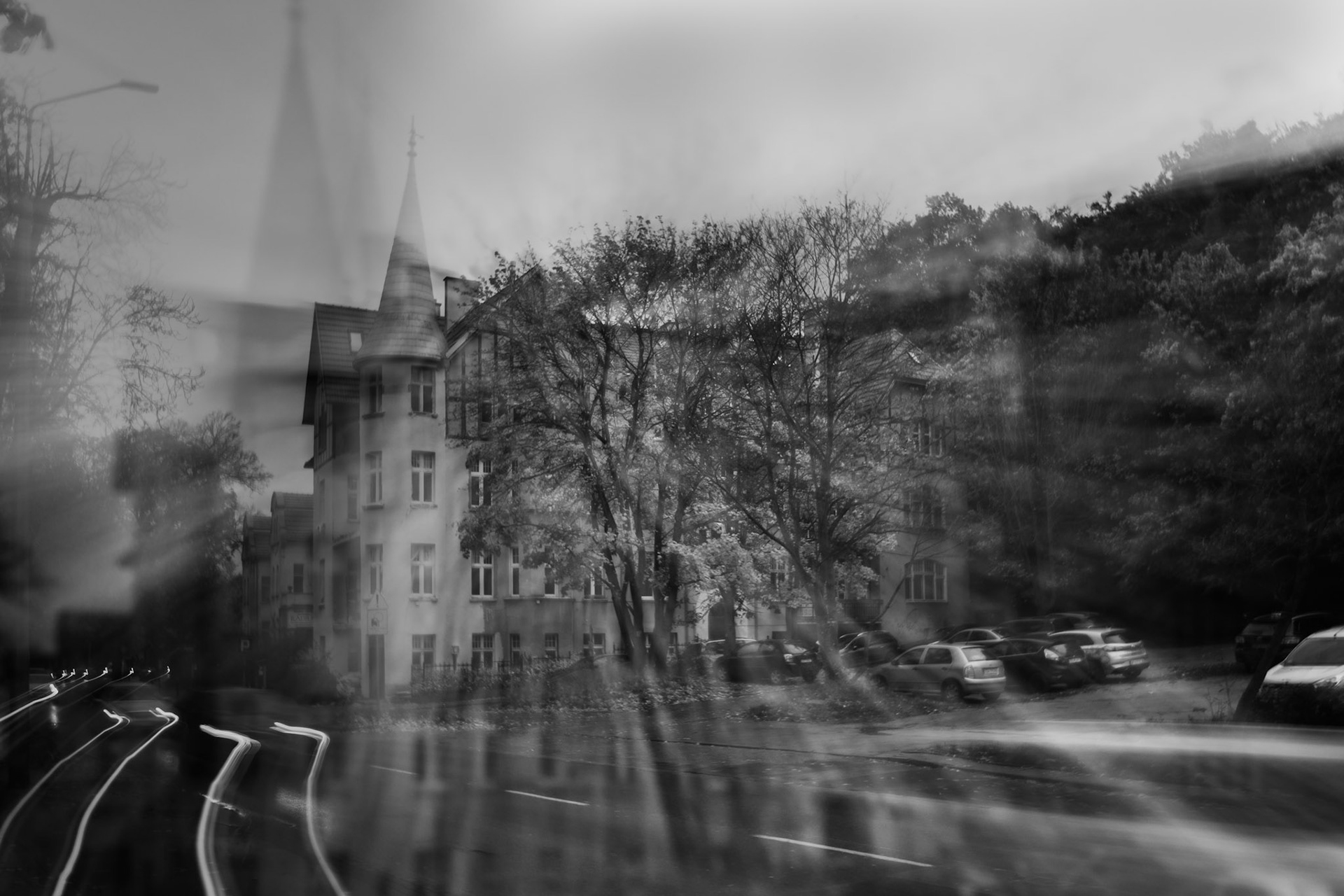 Black and white photo of an old building with a pointed tower, surrounded by trees and parked cars, captured with motion blur on a rainy street with ICM technique.