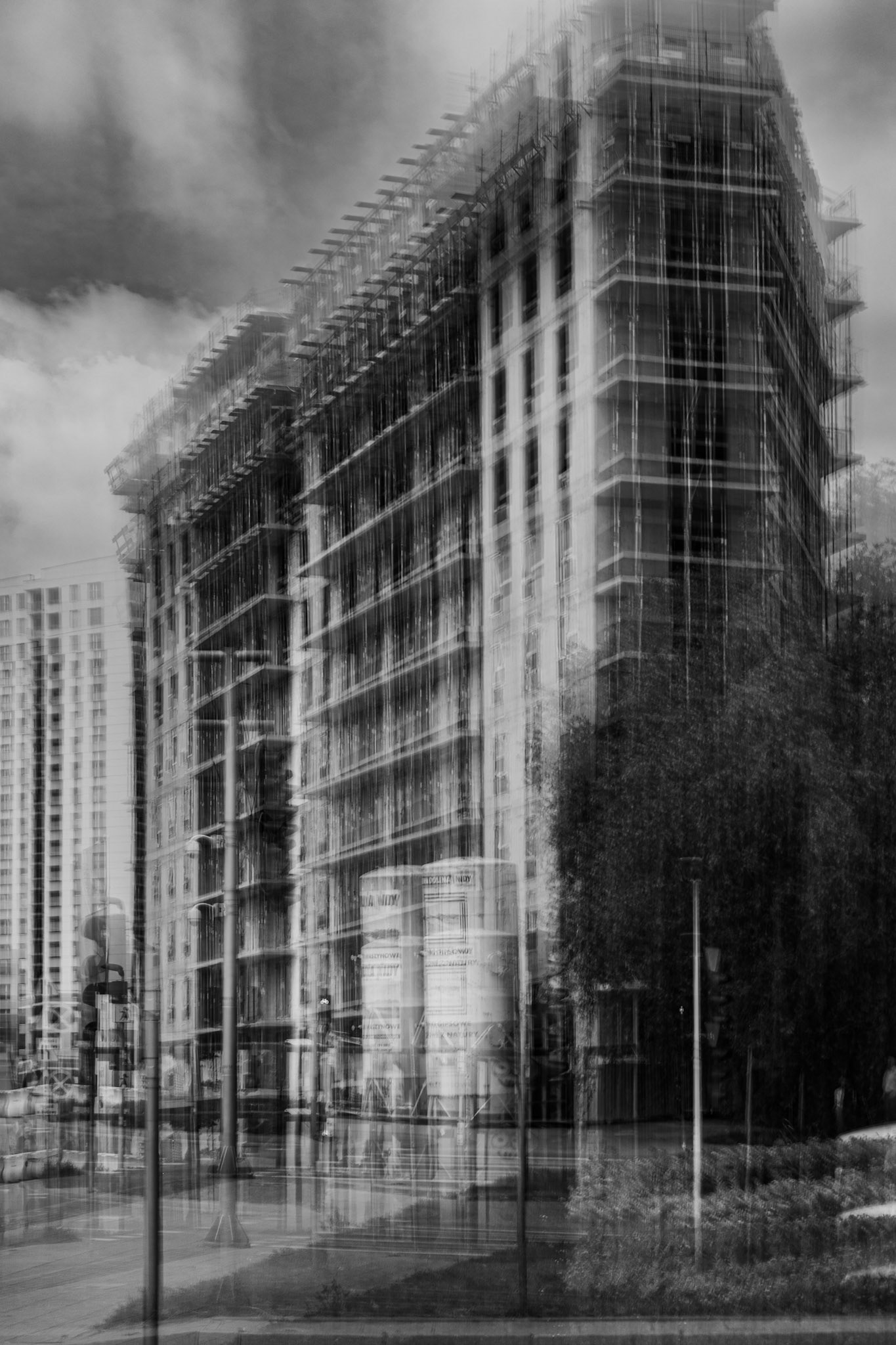 Black-and-white long-exposure photograph of a multi-story building under construction in an urban setting, featuring scaffolding, motion blur, dramatic cloudy skies, and surrounding city streets with pedestrians and streetlights.