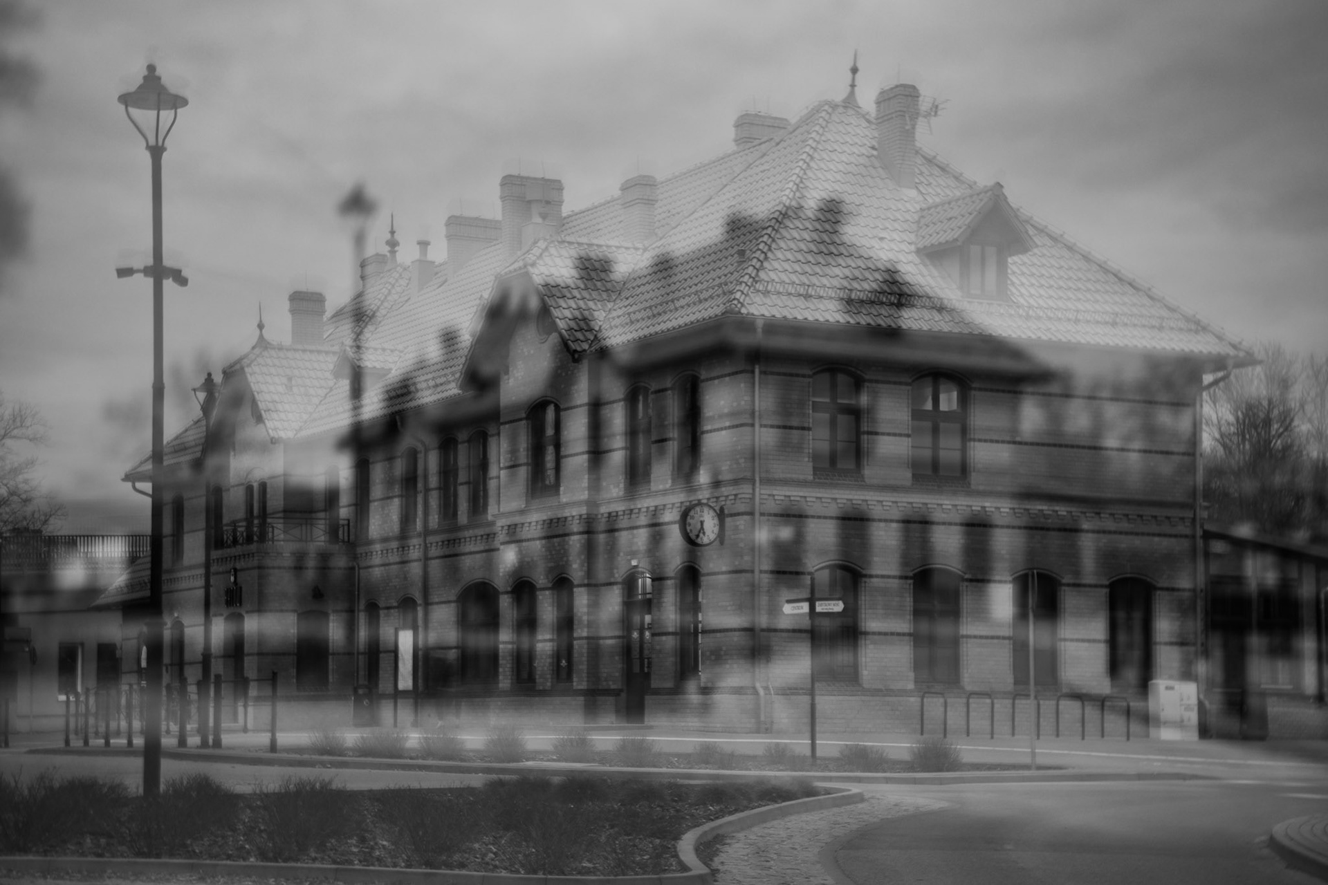 A black and white, double-exposure photograph of a large, two-story brick building, likely a train station, featuring multiple chimneys and a gabled roof. The overlay creates a ghostly, translucent effect, blending the building's facade with a street scene containing lampposts and a paved walkway.