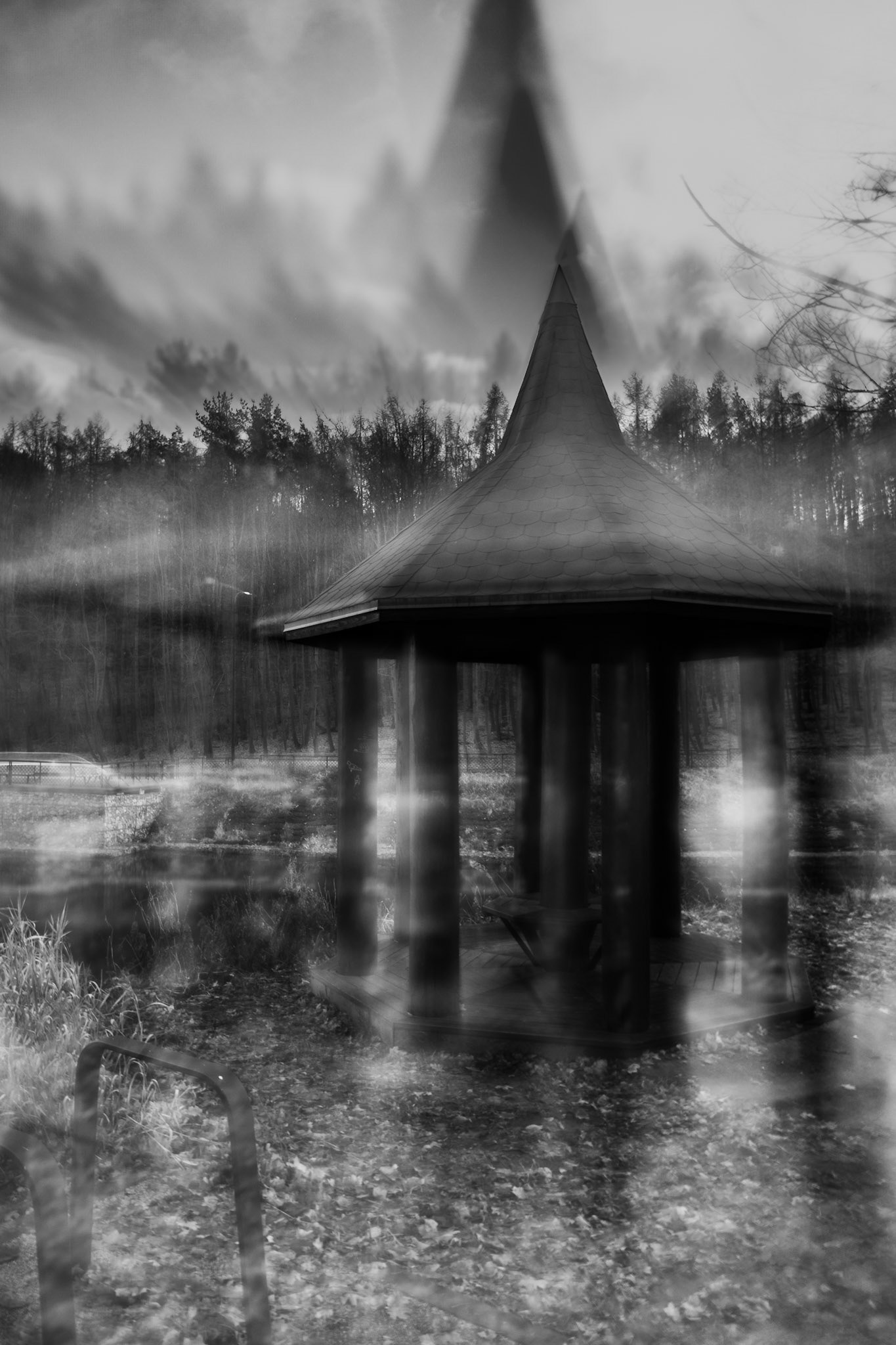 Black-and-white long-exposure photograph of a wooden gazebo in a forest clearing, with motion blur, misty atmosphere, and leaf-covered ground creating an eerie, ghostly mood.