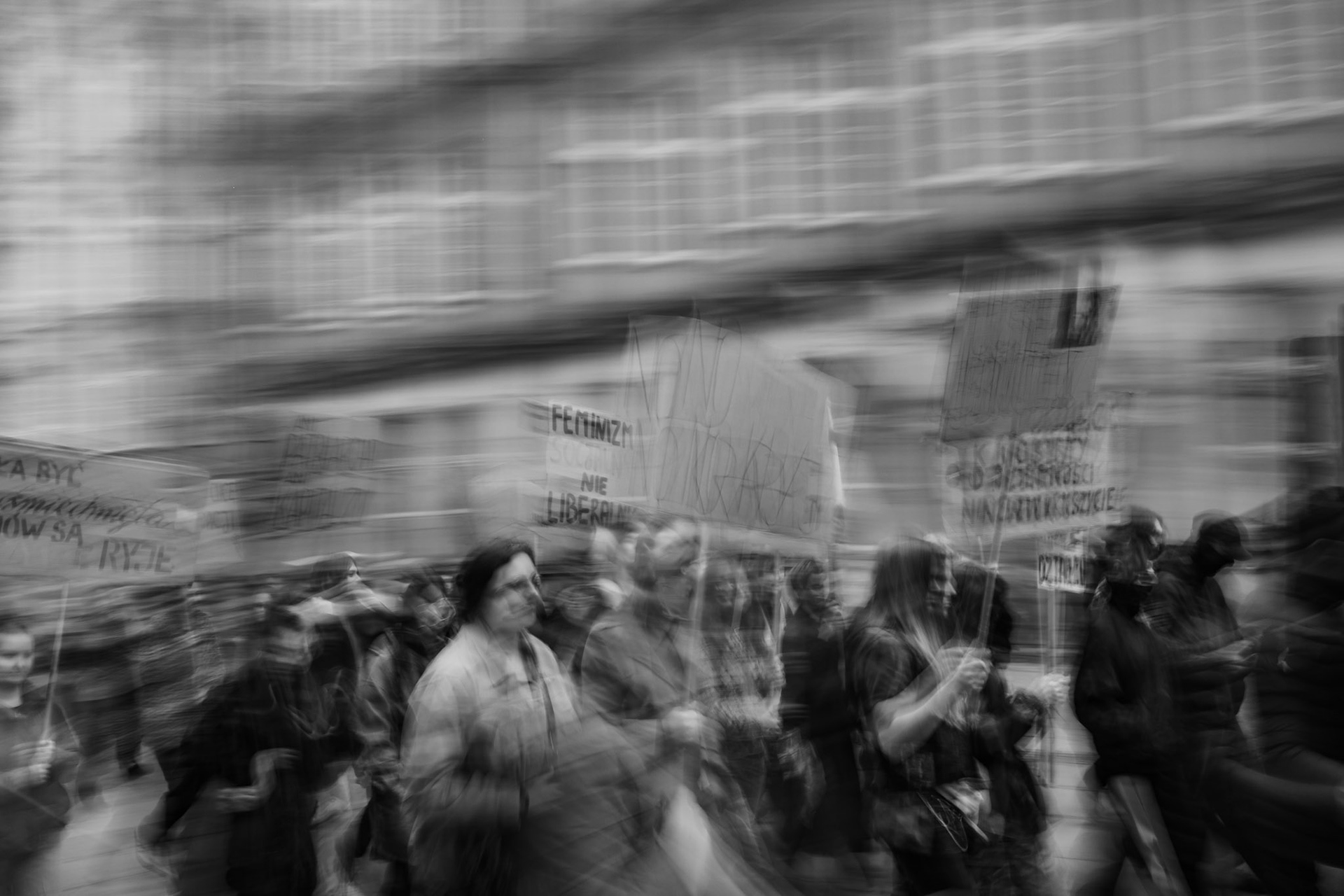 Blurred black-and-white photo of a street protest with a crowd of people marching and holding protest signs, captured with motion blur so the figures and city buildings in the background appear streaked and dynamic.