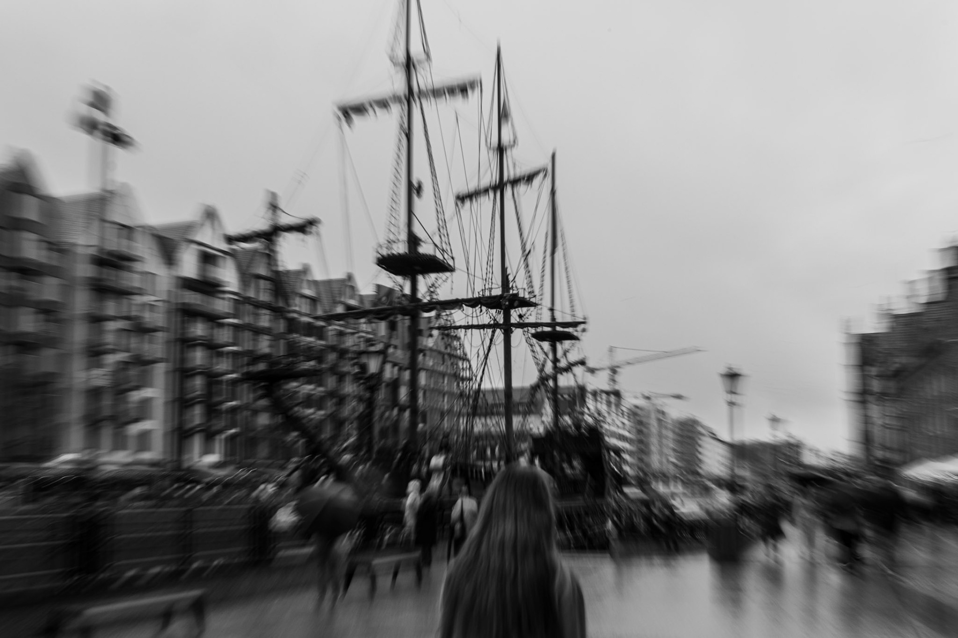 Black and white photography of a paved embankment with people passing by with a reconstruction of a pirate ship in the background