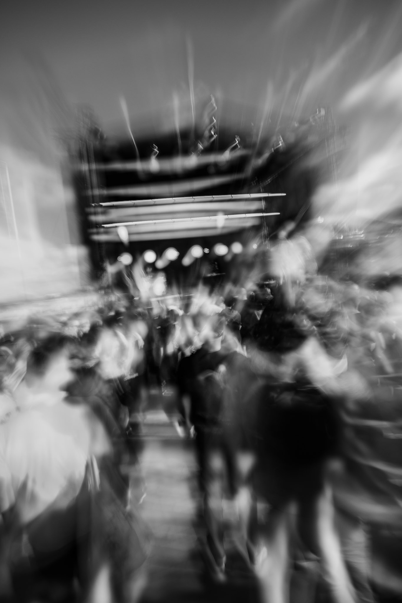 Blurred crowd at an outdoor concert with a brightly lit stage, creating a dynamic motion effect in black and white.